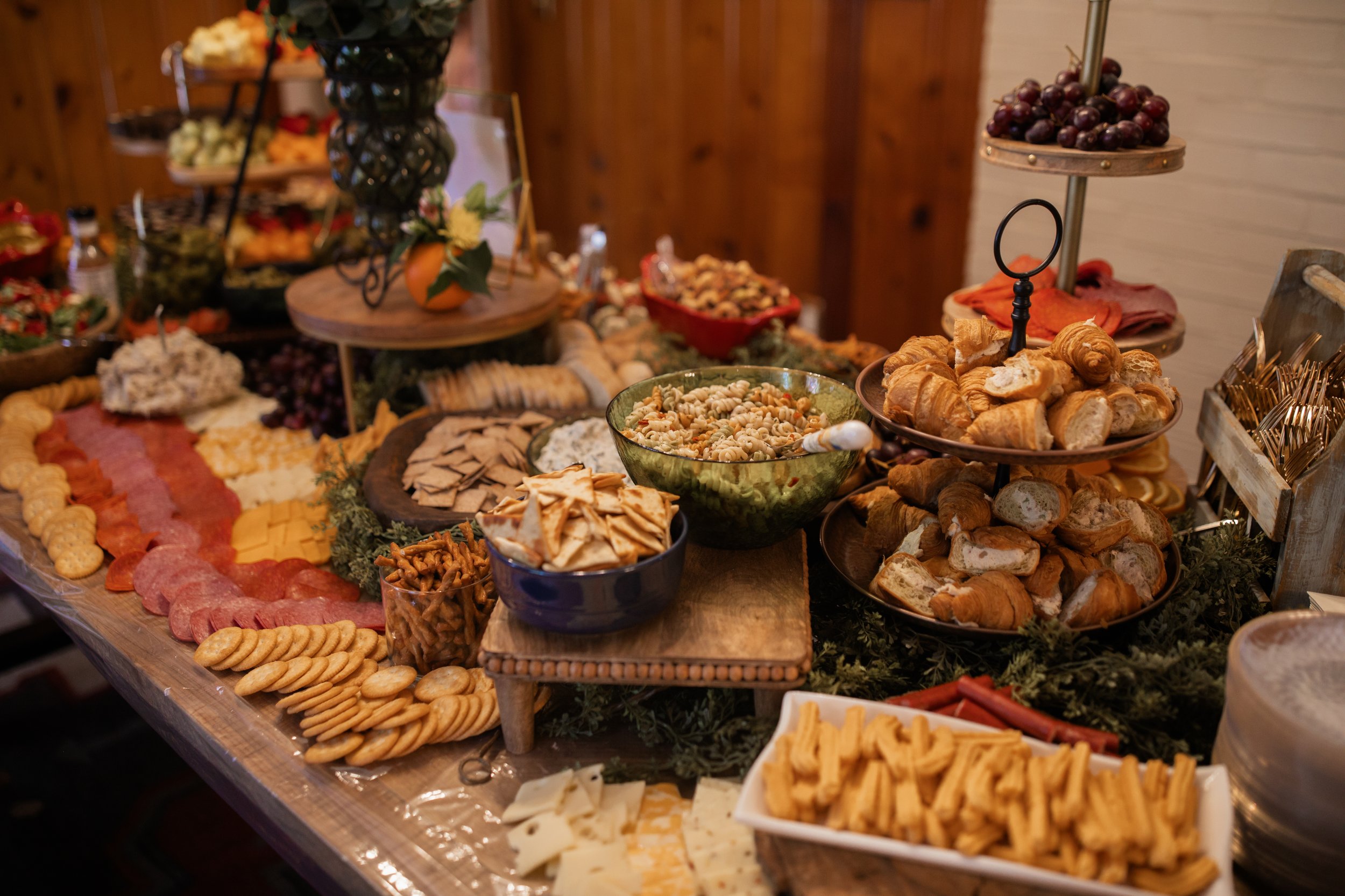 Buffet table with assorted appetizers, including sliced meats, cheeses, crackers, pretzels, croissants, grapes, pasta salad, and various dips.