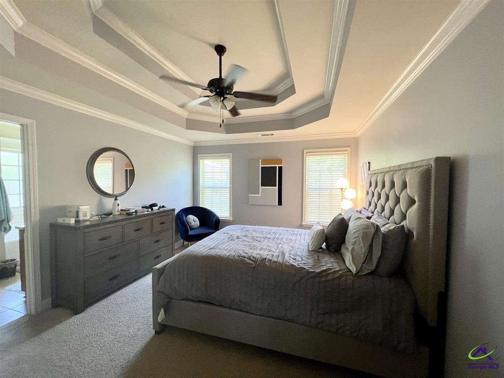 Modern bedroom with a bed, tufted headboard, dresser with a mirror, a chair, artwork, and windows with blinds. Ceiling fan above with tray ceiling design.