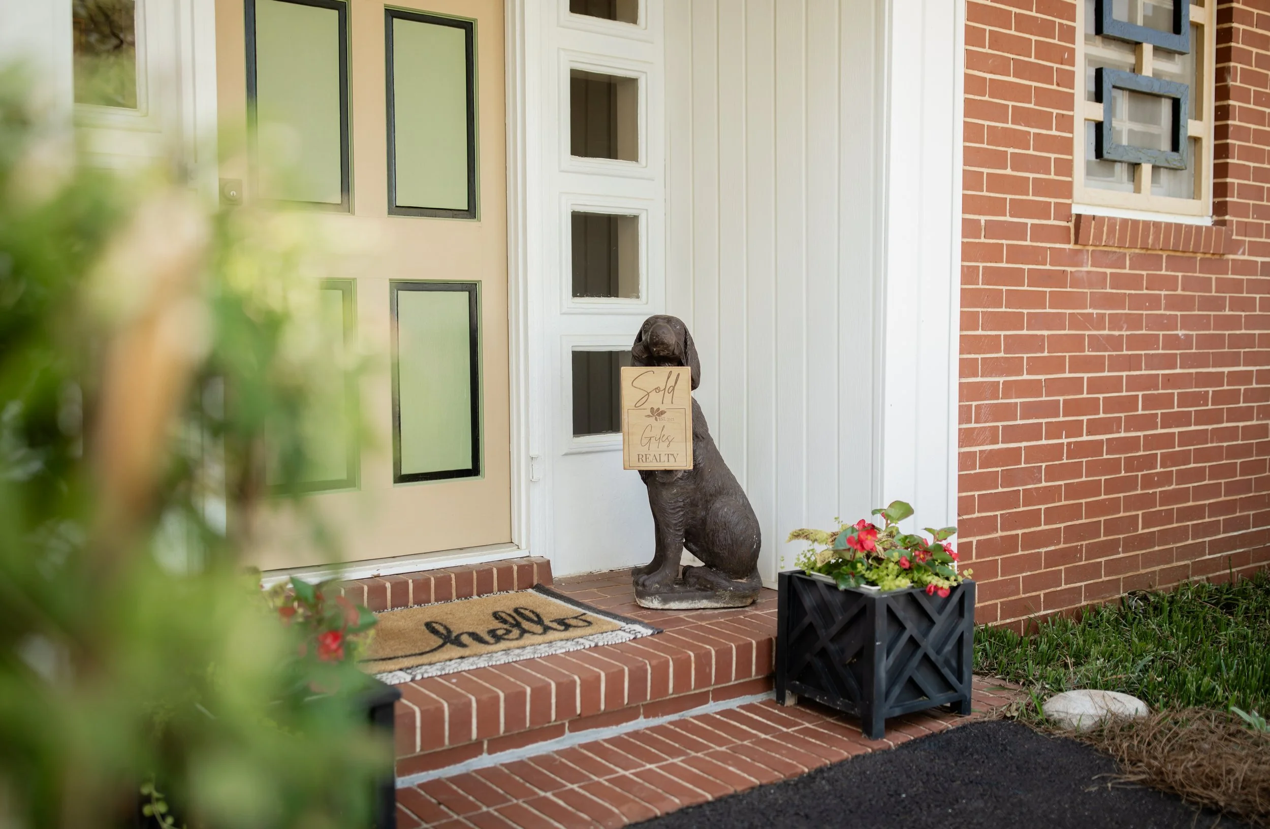 Front porch with a tan front door, black-bordered panels, a dog statue holding a 'Sold' sign, doormat, flower pots, red brick steps, and walls.