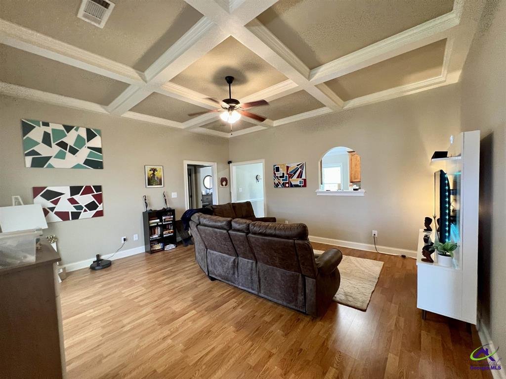 Living room with a brown sectional sofa, wooden floors, coffered ceiling, and colorful abstract wall art. A ceiling fan is centered above the room, and a flat-screen TV is mounted on a white entertainment unit.