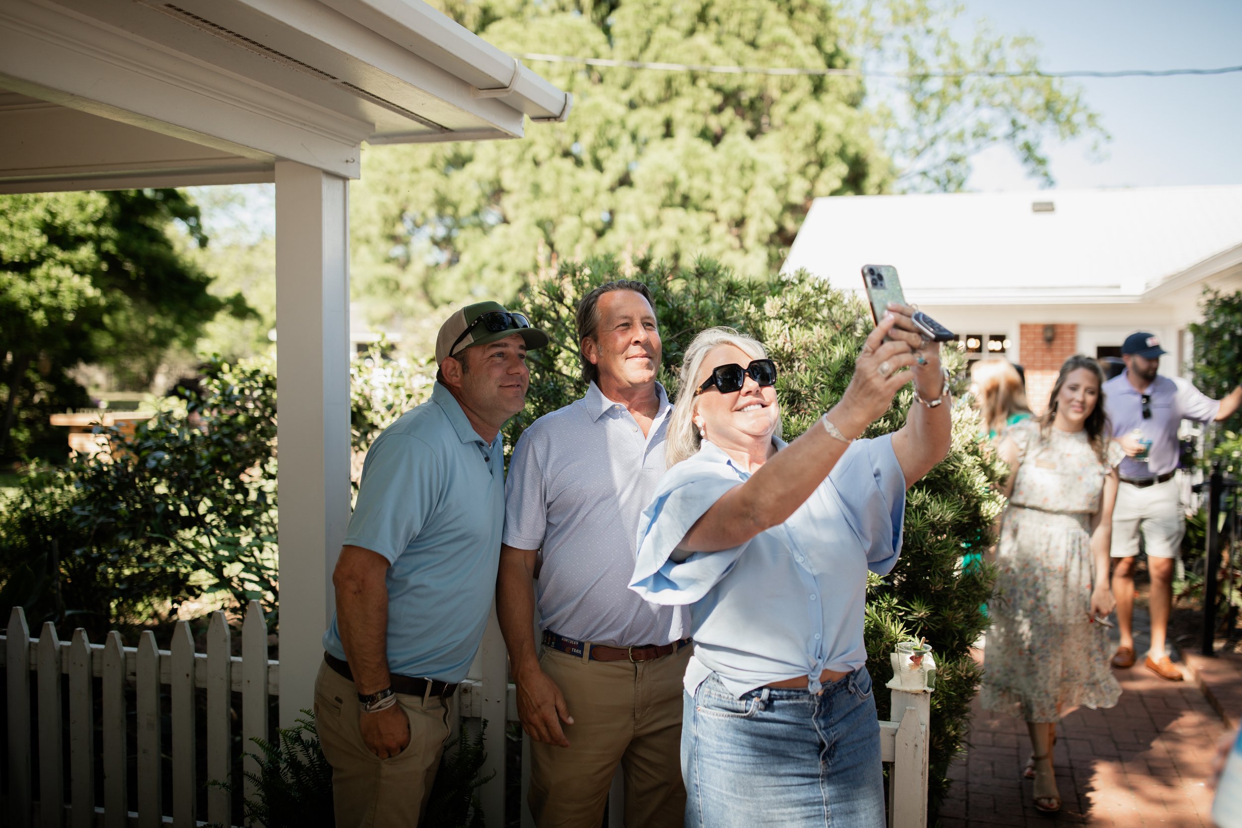 Three people taking a selfie outdoors near a white picket fence, with others socializing in the background.