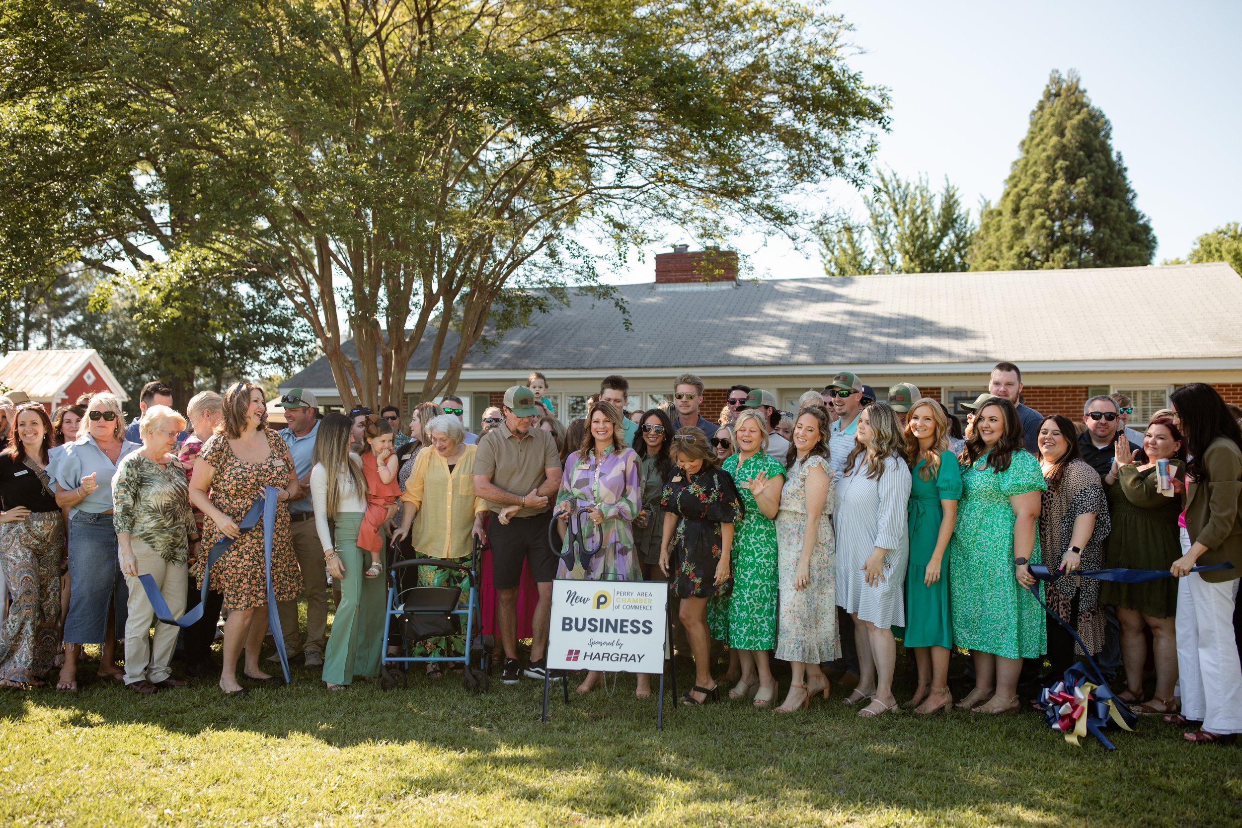 Group of diverse people gathered outdoors for a business event near a building, standing by a sign for Perry Area Chamber of Commerce and Hargray, with a blue ribbon stretched out, indicating a celebration or ribbon-cutting ceremony.