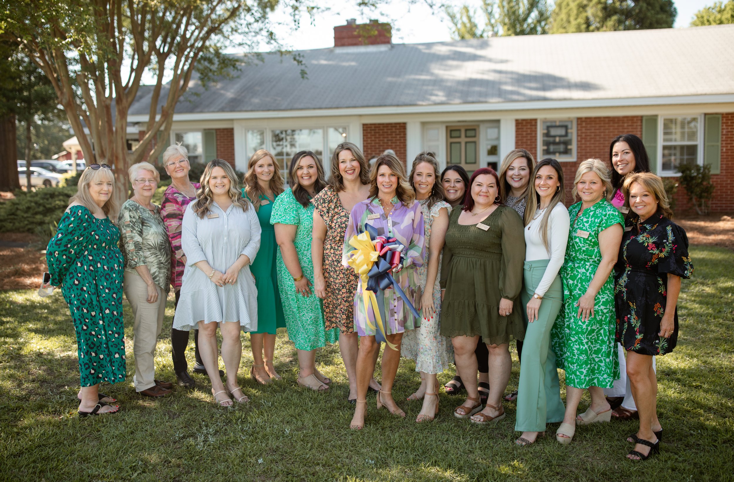 Group of women standing on grass outside a brick building, some wearing green dresses, one holding a bouquet with ribbons.