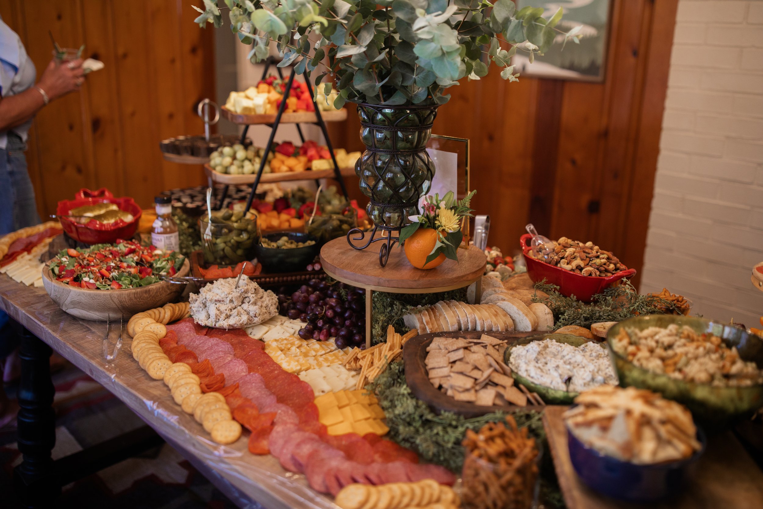 A table set with a variety of foods including a cheese and charcuterie board, bowls of salad, crackers, nuts, and fruit, with a decorative floral centerpiece.