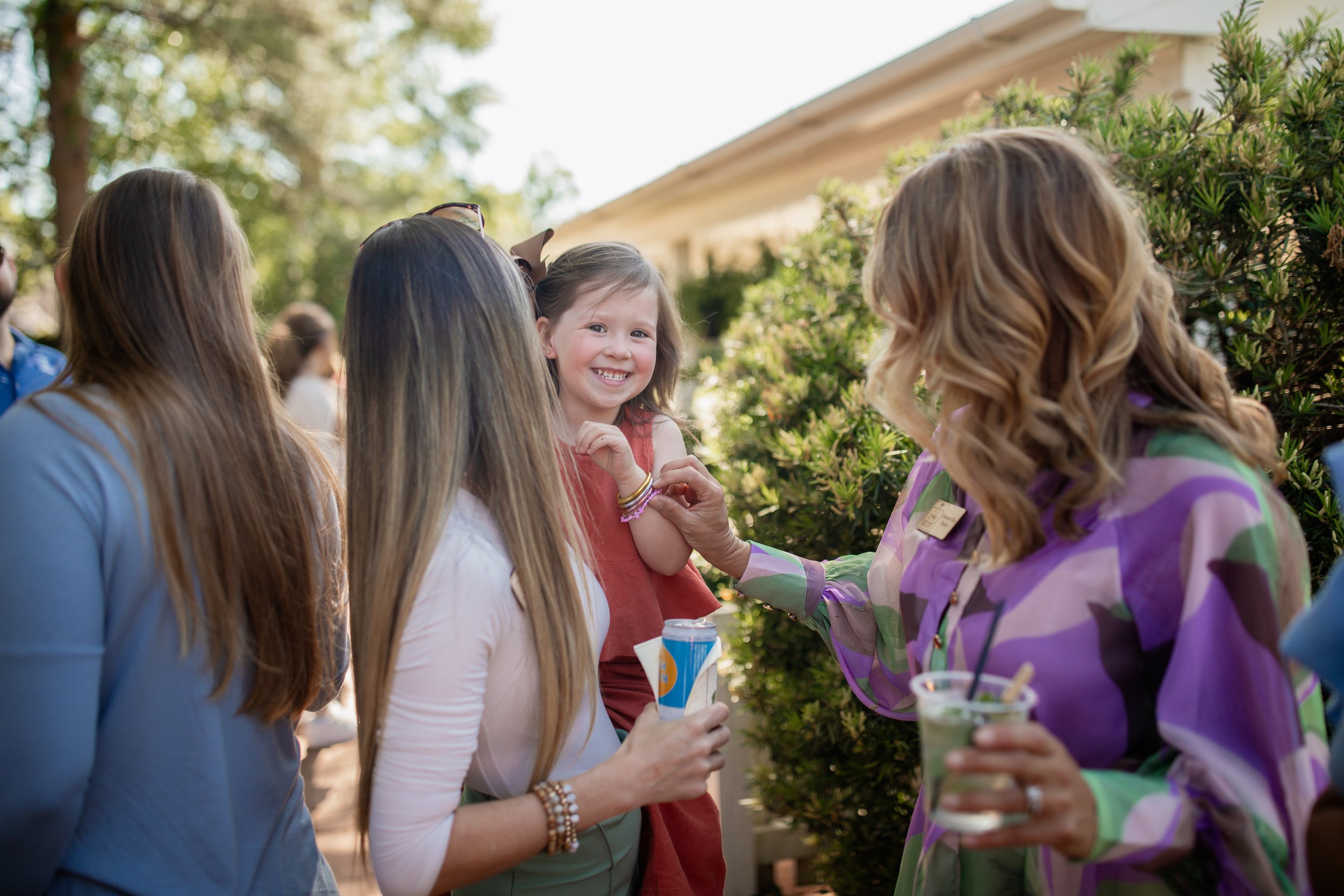 A group of women and a smiling child at an outdoor gathering, surrounded by greenery. One woman is holding a drink, and another is holding a beverage can.