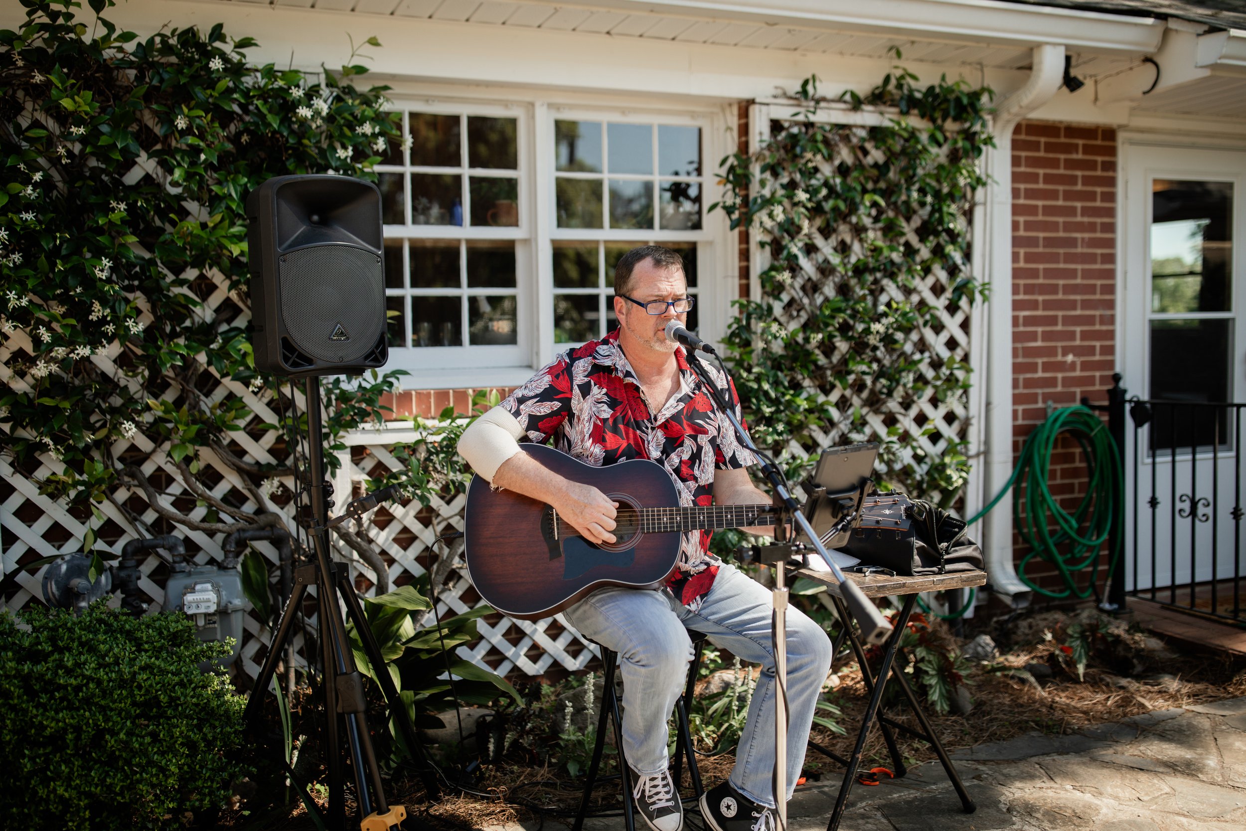 Man playing guitar and singing outdoors with a microphone, wearing a floral shirt and jeans, seated near a speaker and music stand. Background features lattice, greenery, and brick wall.