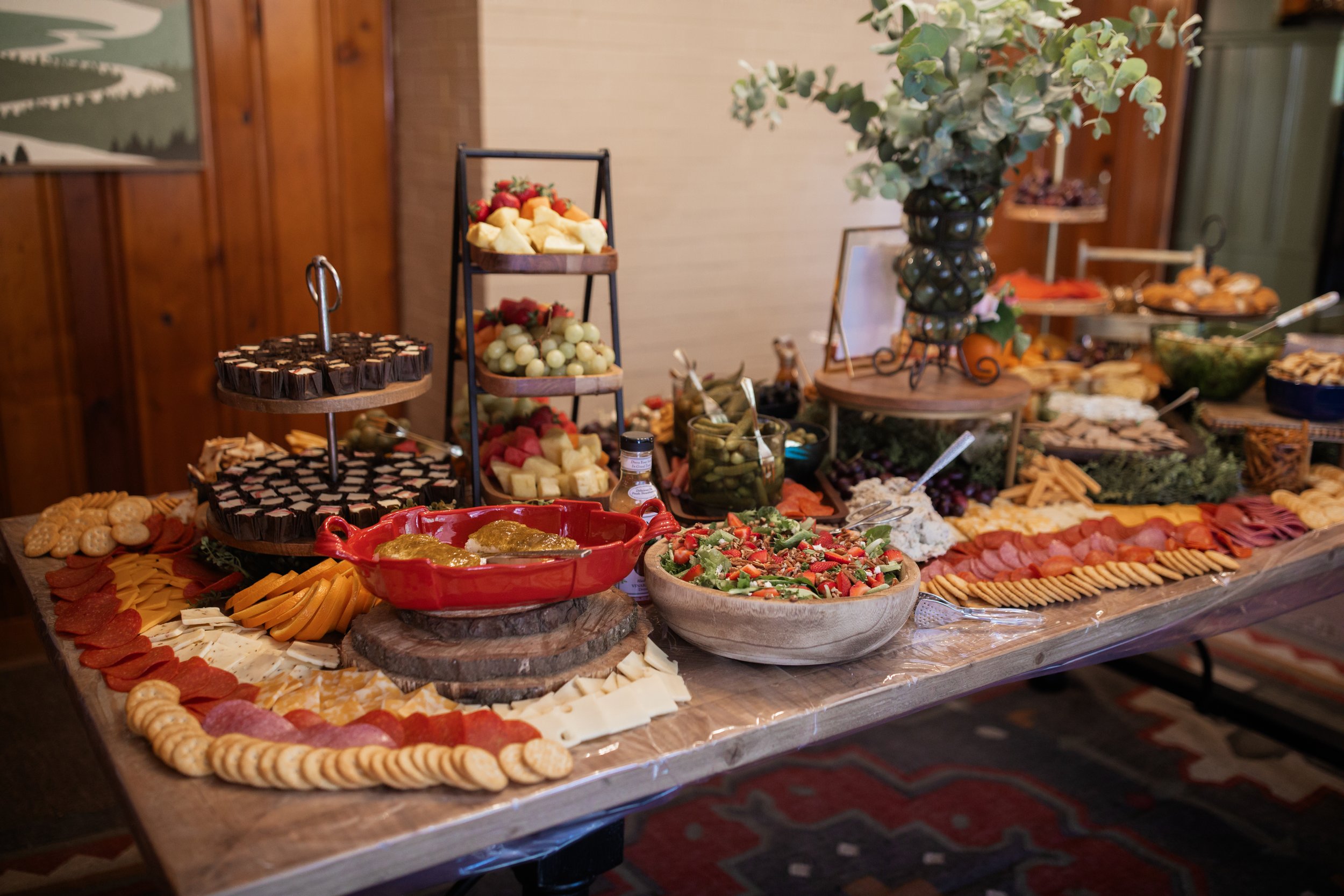 A buffet table featuring an assortment of food, including a variety of cheeses, sliced meats, crackers, salads, pickles, fruit, brownies, and decorative greenery.