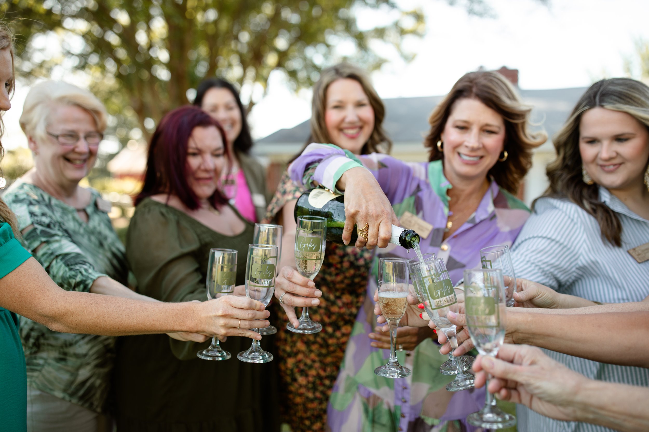 A group of women smiling and holding champagne glasses while one woman pours champagne into the glasses, outdoors with trees in the background.