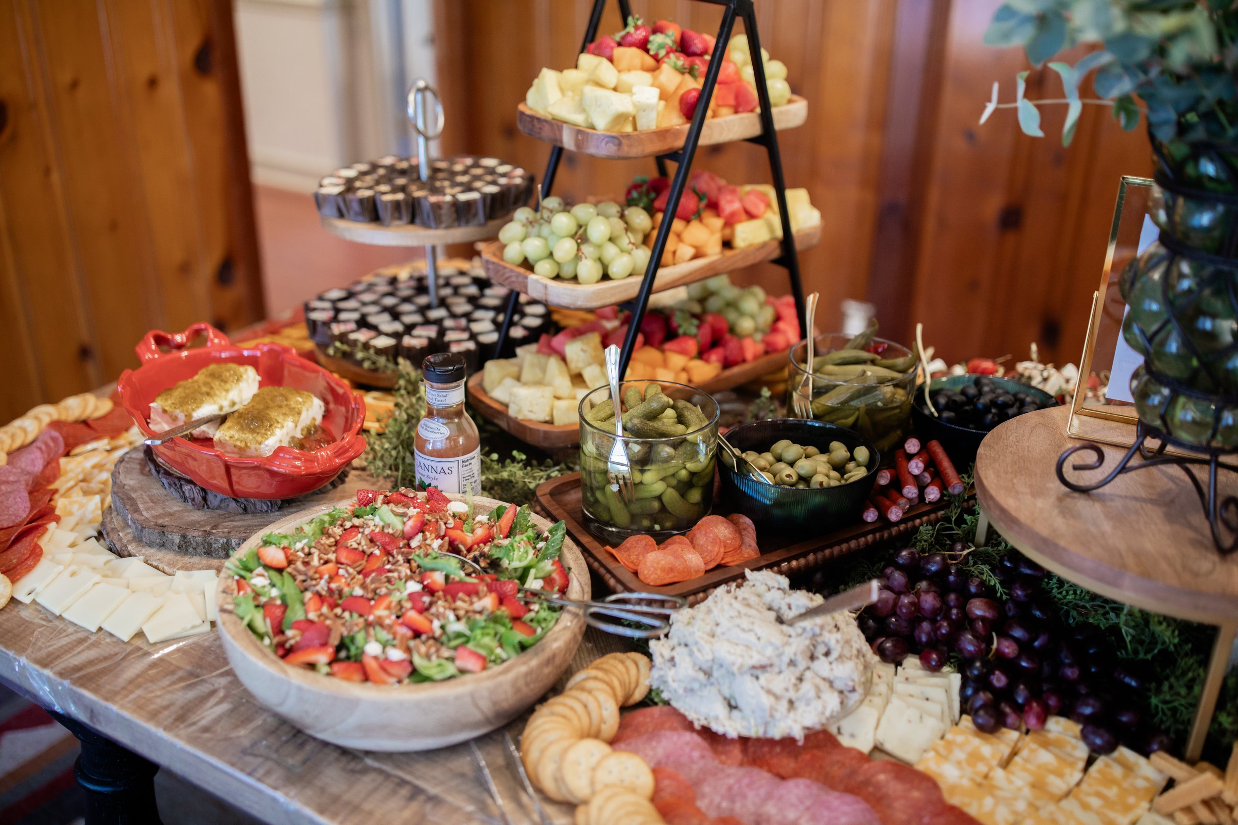 Charcuterie table with assorted cheeses, meats, crackers, fruits, pickles, olives, and salad in a wooden bowl on display.