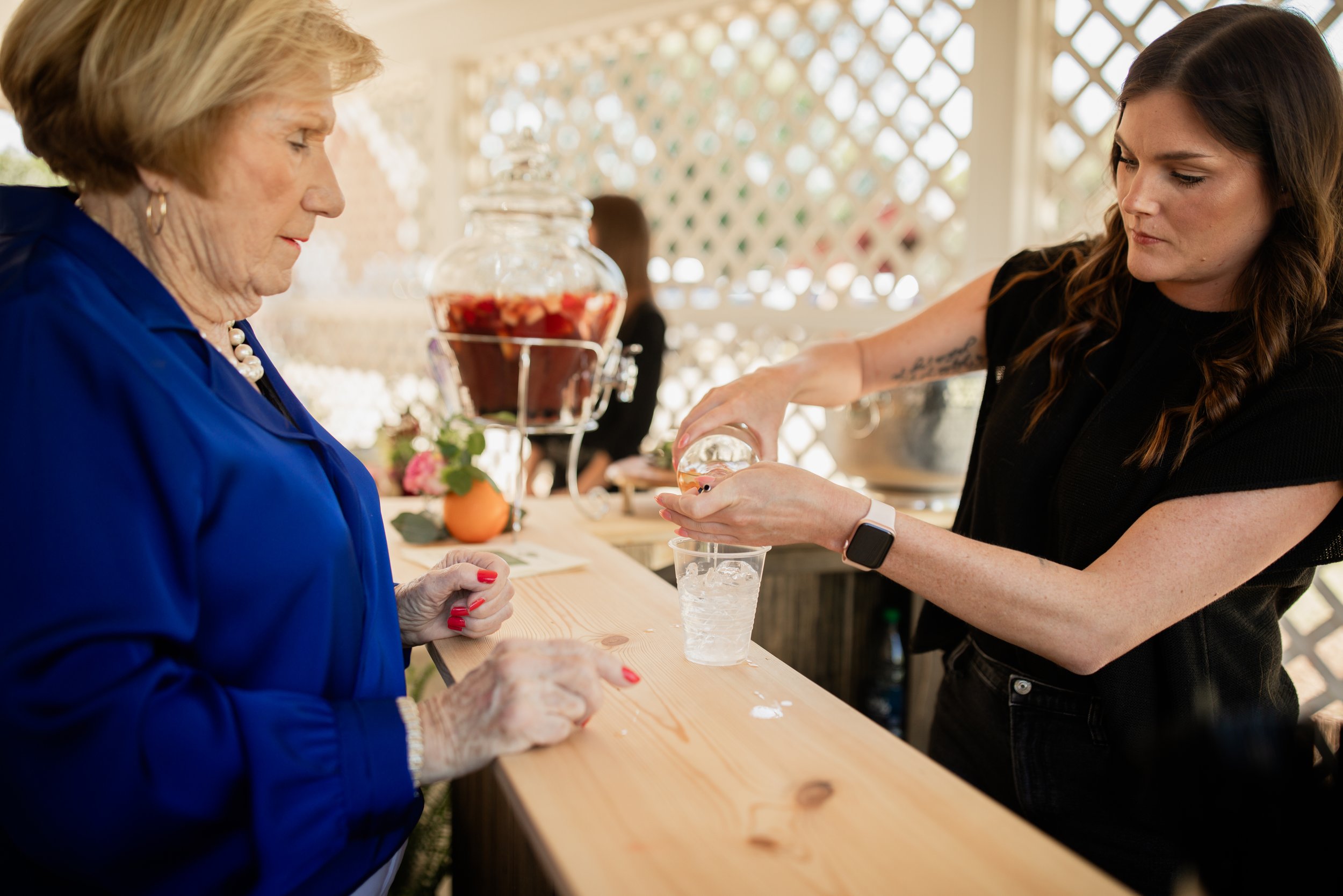 Woman in blue jacket at a drink stand, another woman pouring beverage into a cup with ice, large jar of fruit-infused drink on counter.