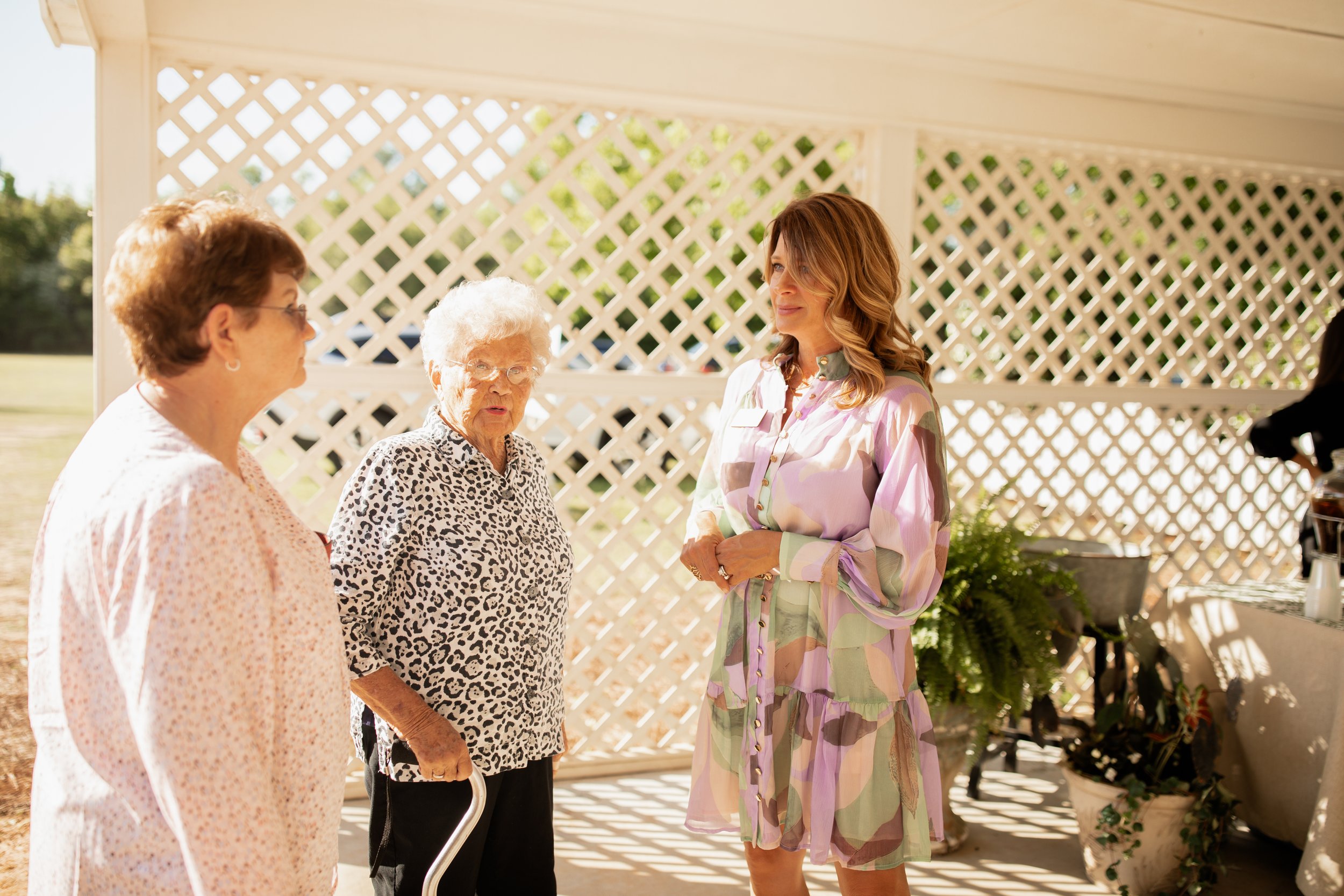 Three women conversing under a gazebo with white lattice walls; one elderly woman using a cane, another in a pink blouse, and a third in a colorful dress.
