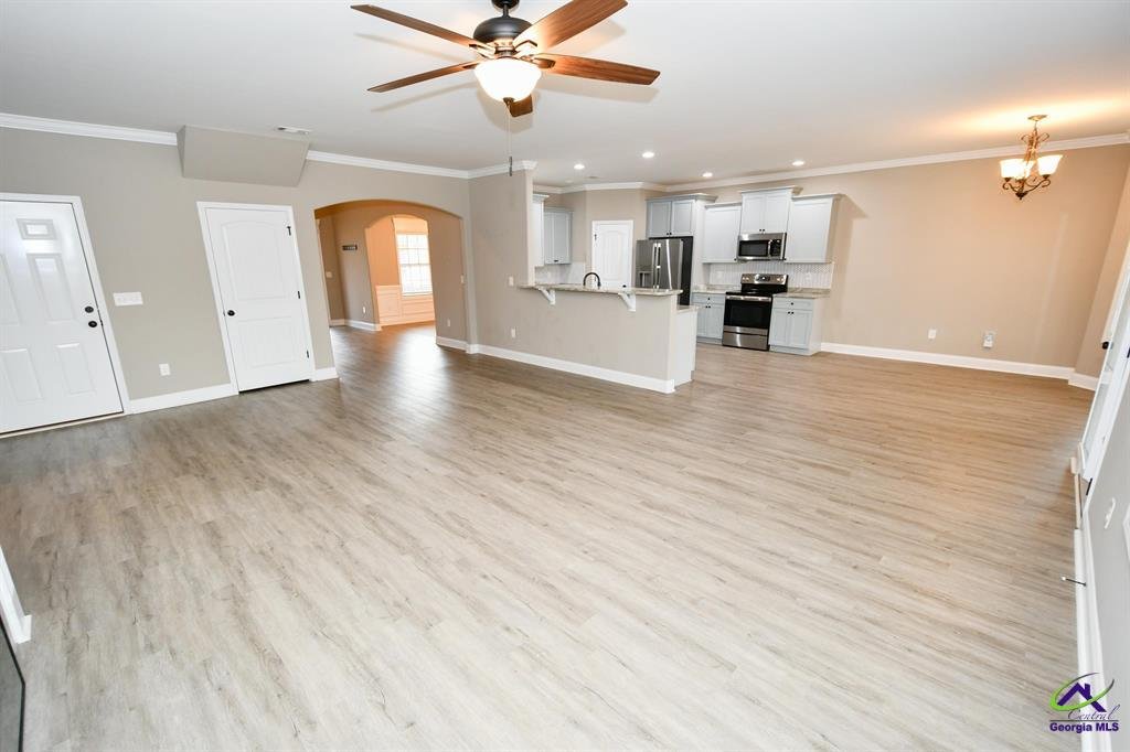 Spacious open-concept living area with light hardwood flooring, featuring a ceiling fan, kitchen island with sink, stainless steel appliances, white cabinetry, and beige walls. Recessed lighting and a chandelier enhance the modern look. A hallway lea