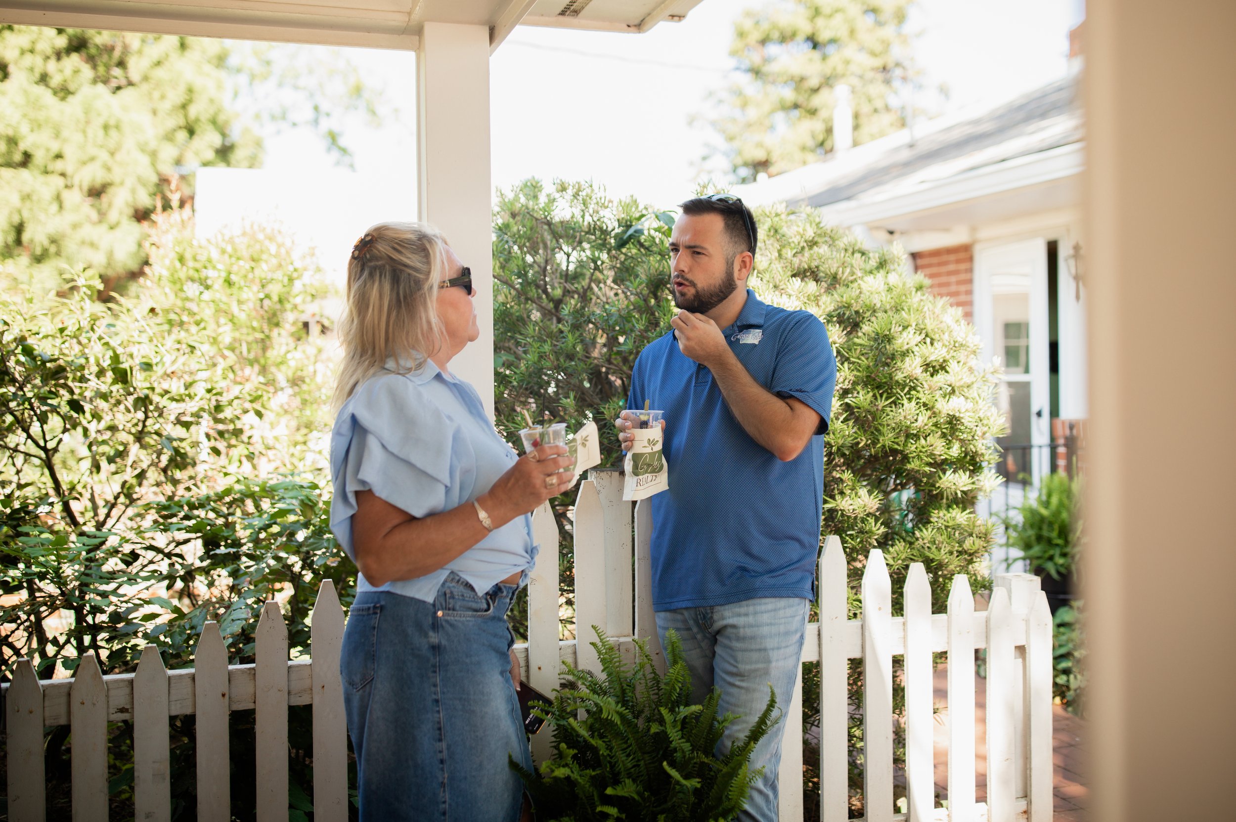 Two people talking outdoors near a white picket fence, holding drinks, with greenery in the background.