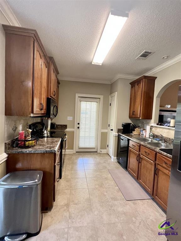 Modern kitchen with wooden cabinets, stainless steel appliances, and tile flooring. Features a central fluorescent light, a trash can, and a mat on the floor. A door with glass panes leads outside.