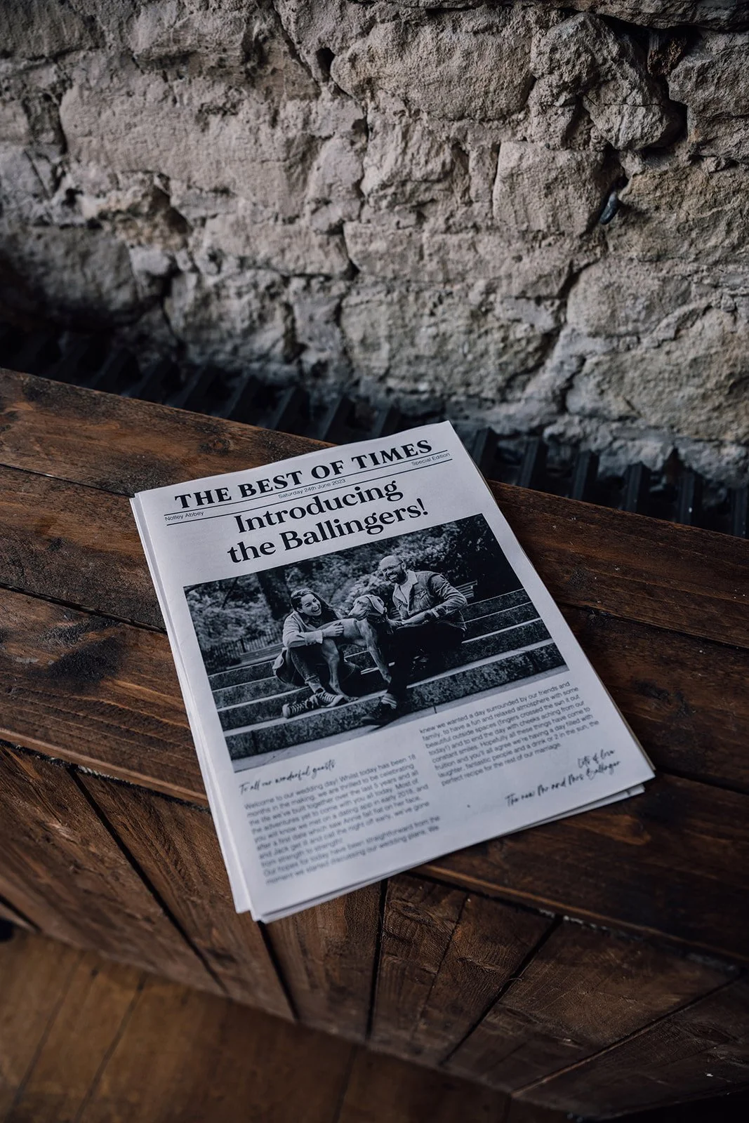 A black and white newspaper titled 'Introducing the Ballingers!' resting on a wooden surface, with a stone wall in the background.