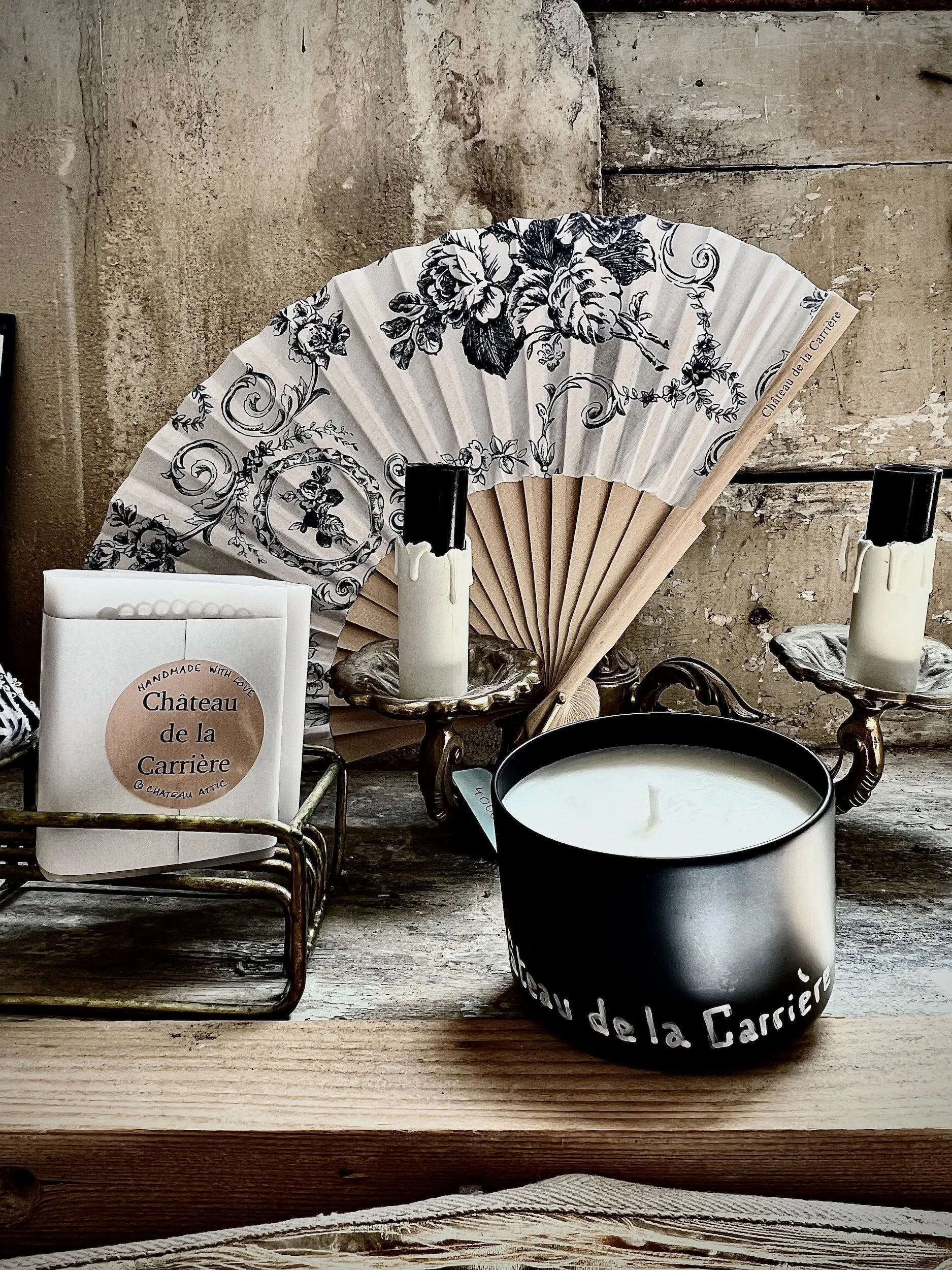 Decorative arrangement including a black and white fan with floral patterns, candles in holders, a black candle with 'Château de la Carrière' written on it, and a small box with a label that reads 'Château de la Carrière' on a rustic wooden surface.