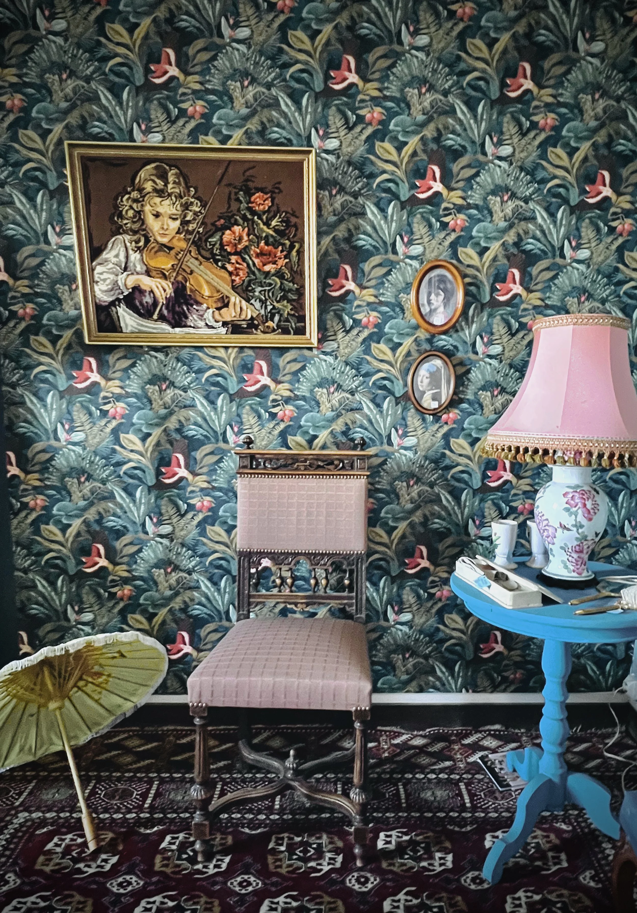 A vintage interior scene with floral wallpaper, an empty antique chair, a pink table lamp, and small framed portraits on the wall. Decorative items include an umbrella on the floor and a tray with utensils on the table.