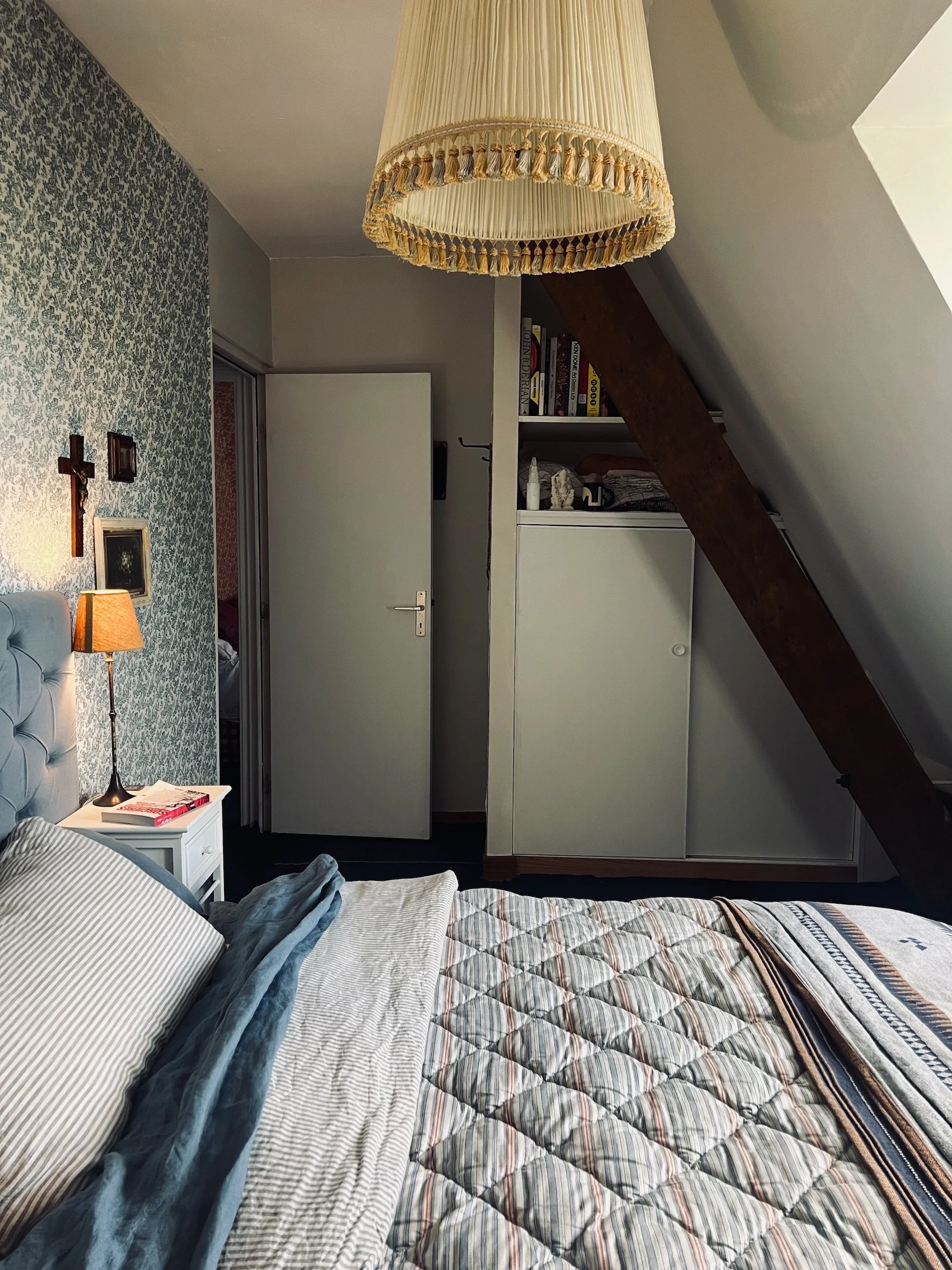 The Attic, A bedroom with a bed, bedside table, lamp, and hanging ceiling lamps, decorated with crosses and patterned wallpaper, under a sloped ceiling with a wooden beam.