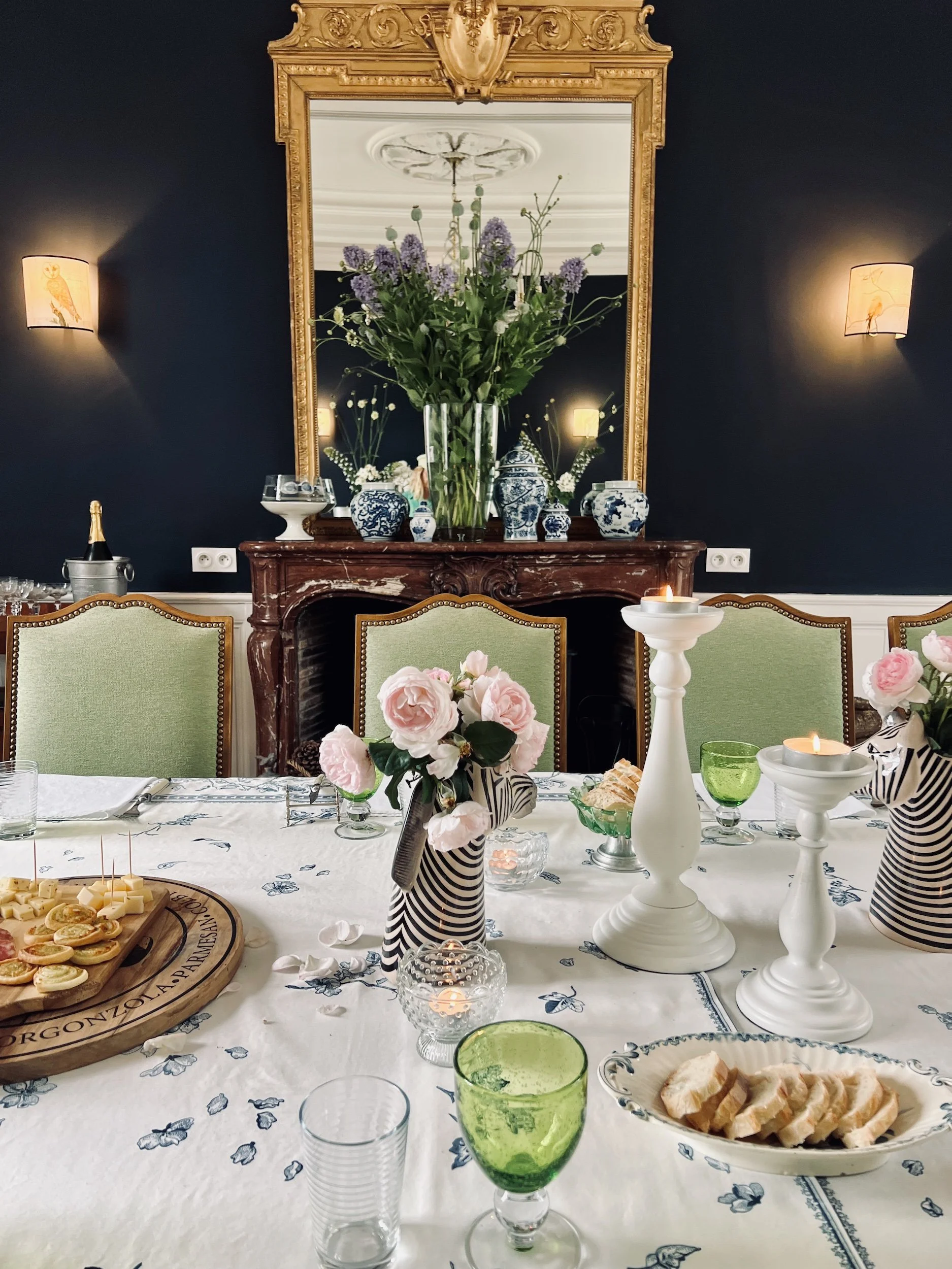 Elegant dining room table setup with floral centerpieces, candles, and assorted snacks, against a dark wall with a large mirror and decorative vases.