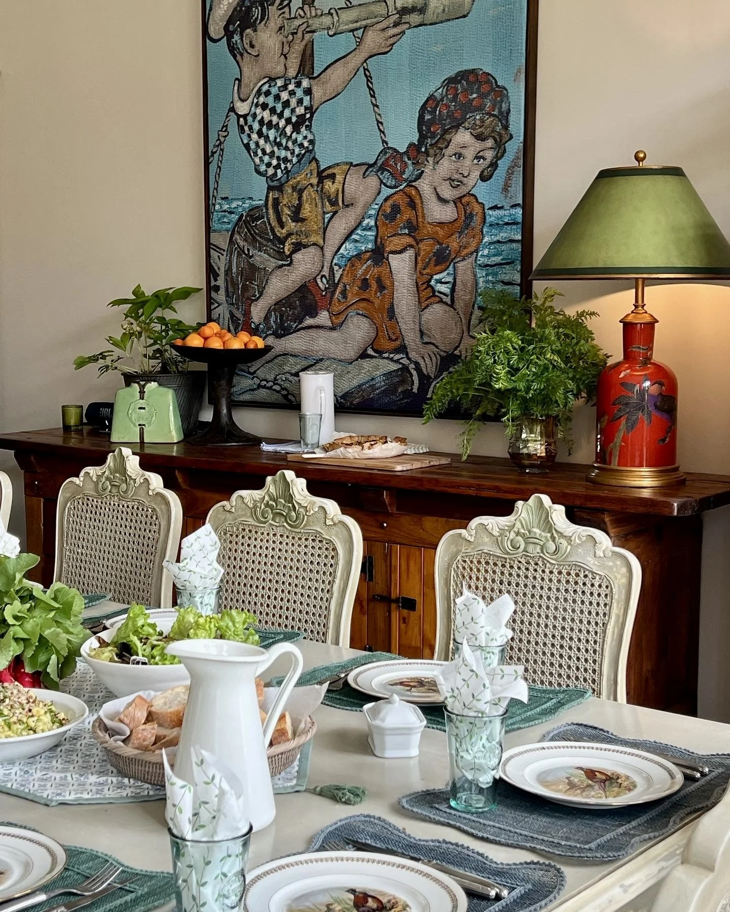 A dining room setup with a table set for a meal, featuring plates with bird illustrations, glasses, utensils, napkins, and a basket of bread. In the background, a wooden sideboard with plants, a bowl of oranges, a lamp with a green shade, and a colorful painting of two children near the water are visible.