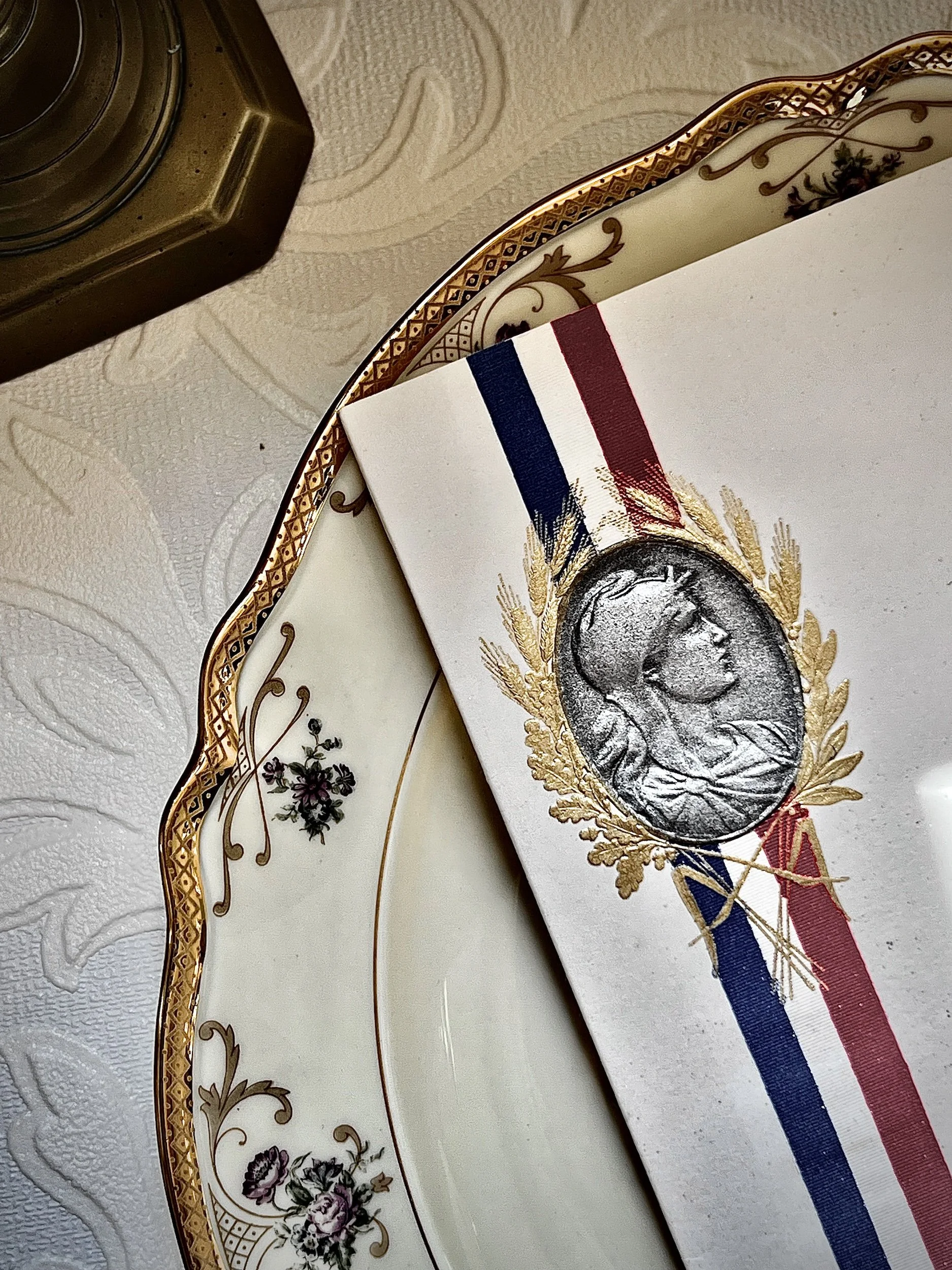 A decorative plate with gold and floral details, a white paper with a medal featuring a woman's profile surrounded by a gold laurel wreath, and a gold-edged glass container on textured white tablecloth.