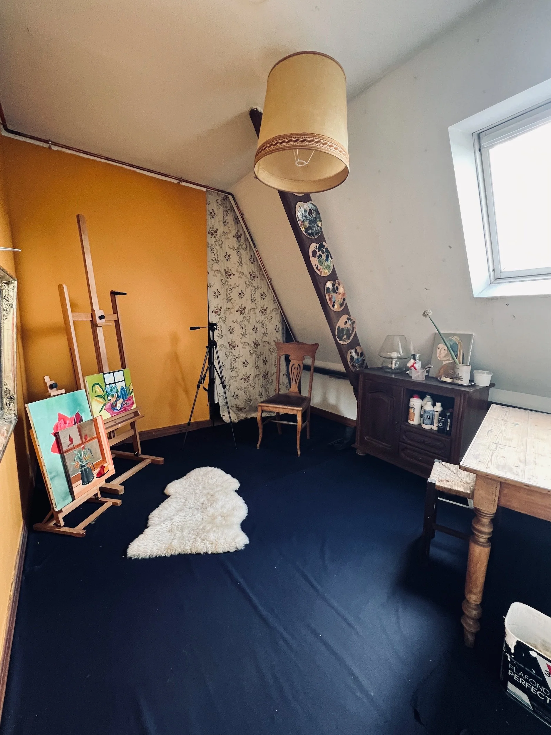 An art studio with yellow and floral wallpaper walls, depicting paintings on easels, a white fluffy rug, a wooden chair, a vintage cabinet, and a table near a window with art supplies.
