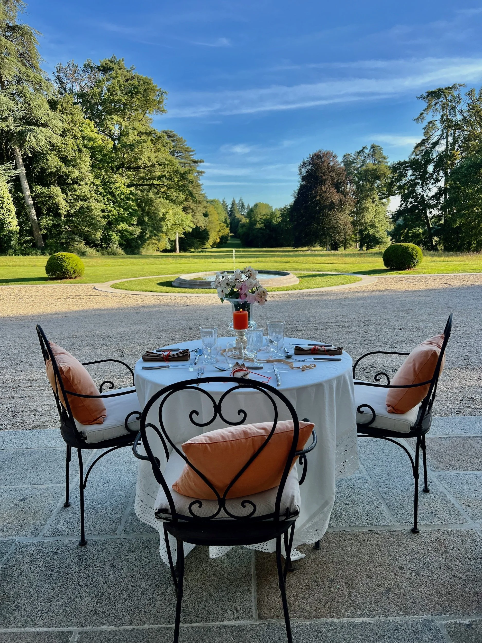An outdoor table set for dining on a patio with a scenic garden view, featuring black wrought iron chairs with peach cushions, a white tablecloth, floral centerpiece, glasses, and table settings, with a fountain and lush trees in the background under a blue sky.