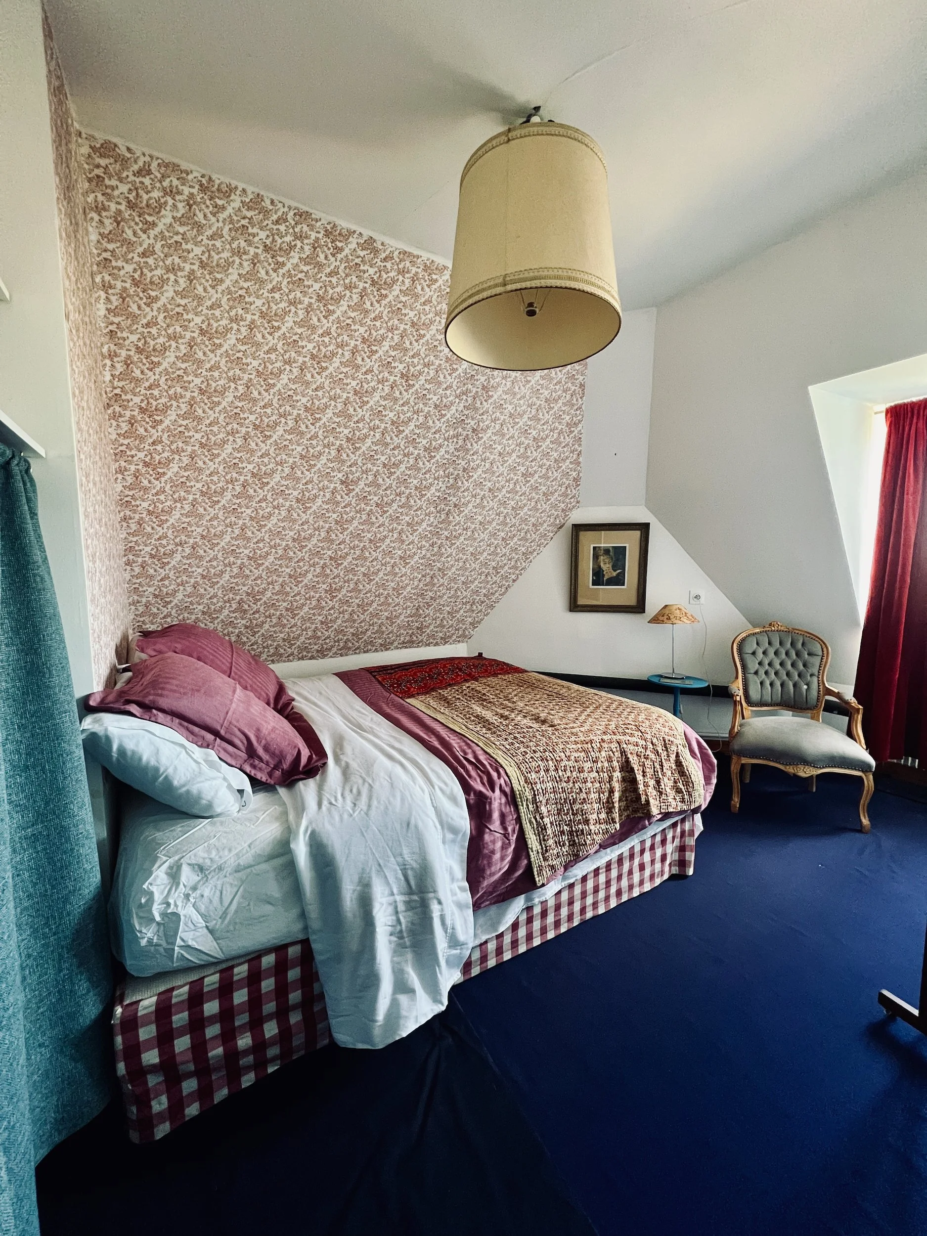 The attic, A bedroom with a bed against a wall with floral wallpaper, a padded armchair near a window with red curtains, and a ceiling-mounted lampshade.