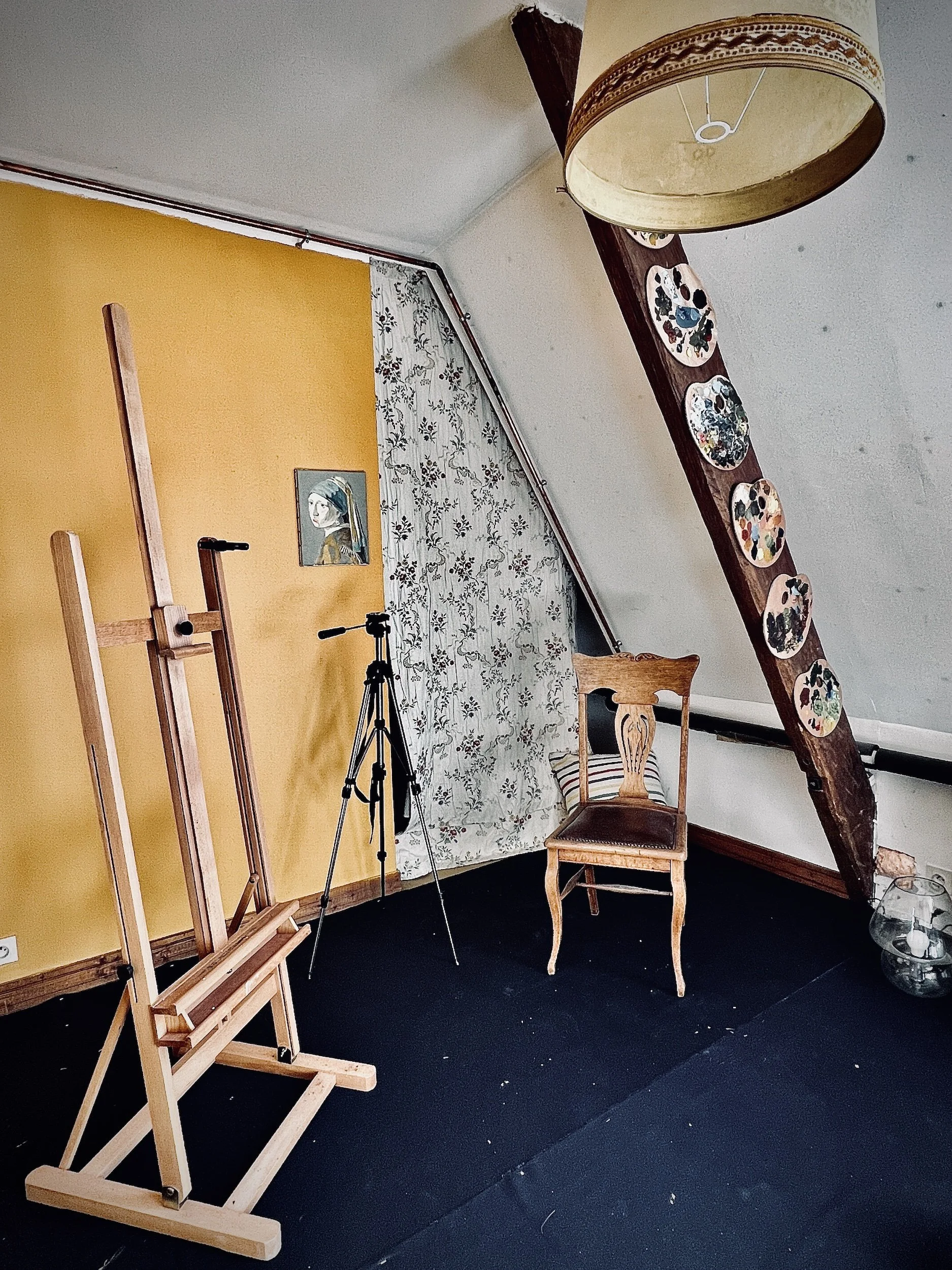 Art studio with a yellow wall, an easel, a camera on a tripod, a floral curtain, a portrait painting, a wooden chair with a striped cushion, and a diagonal wooden shelf with six decorative plates. A large lamp hangs from the ceiling and a glass container with candles is on the floor.