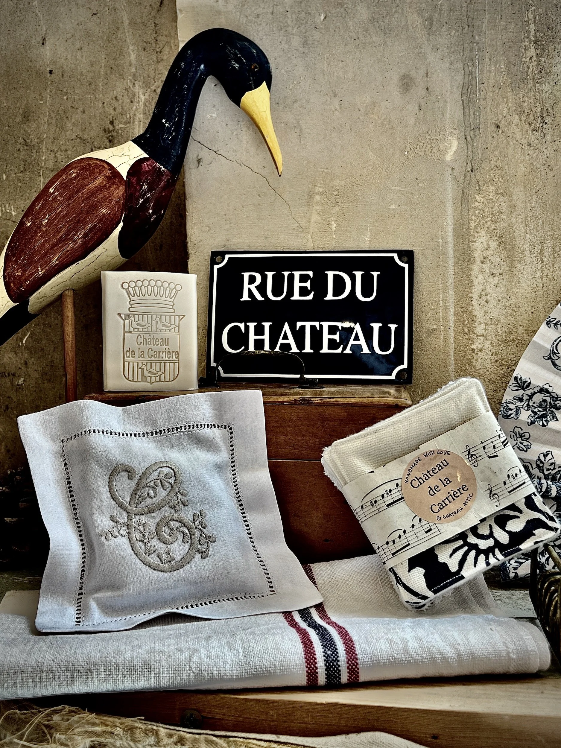 Decorative display with a wooden bird sculpture, a black and white 'Rue du Chateau' street sign, a white embroidered pillow, a small fabric bundle with a 'Made with Love Chateau de la Carriere' label, and a white and black floral patterned cloth on a wooden surface against a rustic wall.