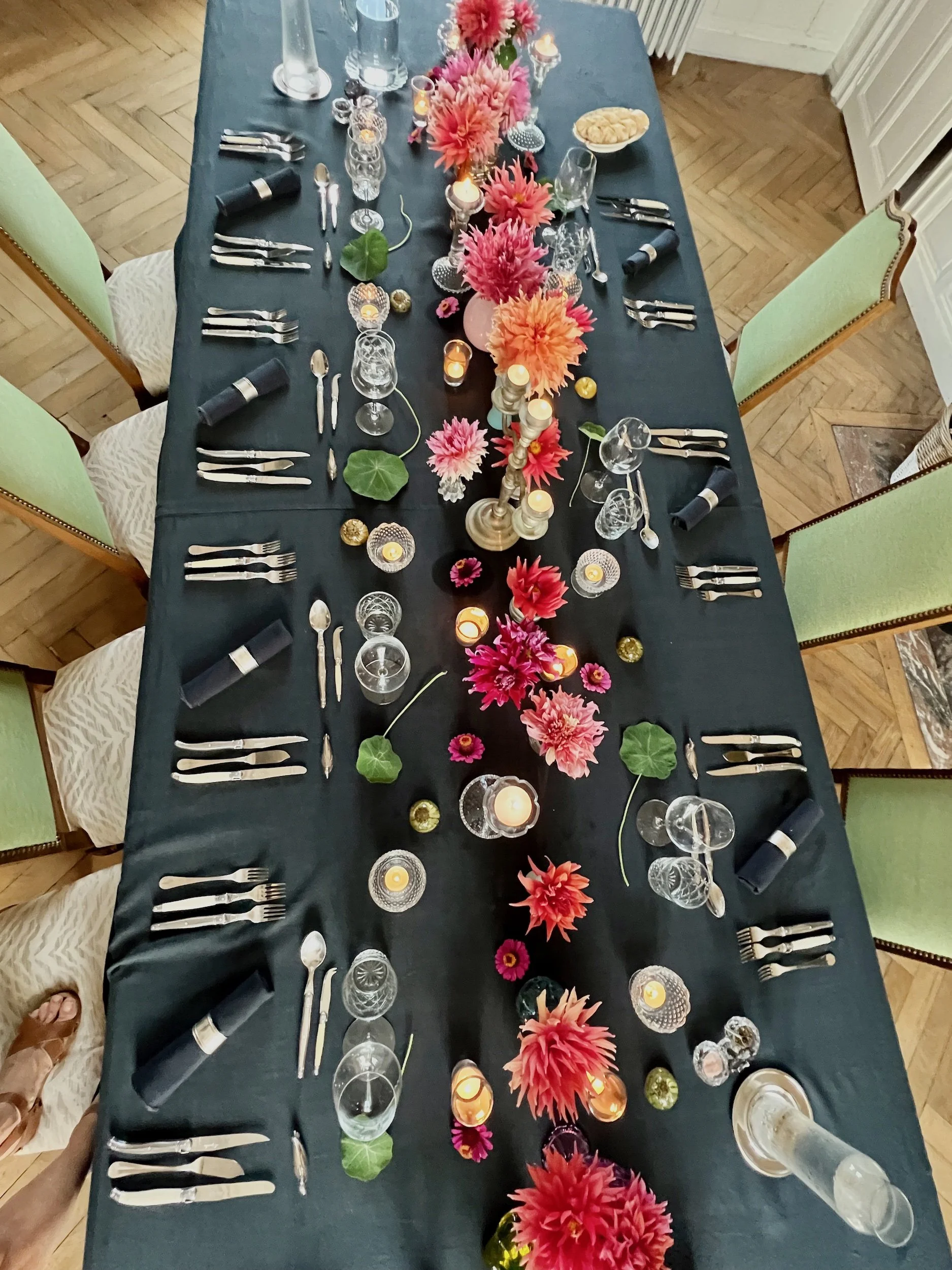 A long dining table decorated with pink and orange flowers, candles, and glassware, set for a meal with green chairs surrounding it.