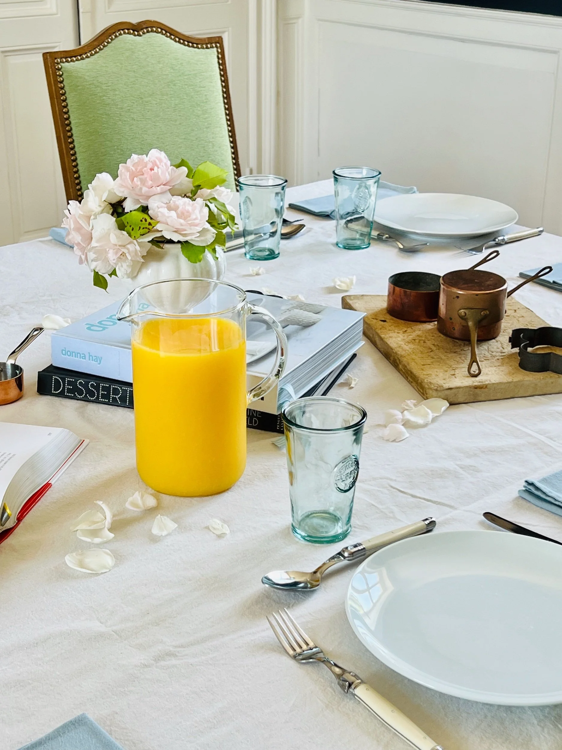 A dining table set with a vase of pink and white flowers, a pitcher of orange juice, blue glassware, white plates, silverware, and some books, with a green chair in the background.