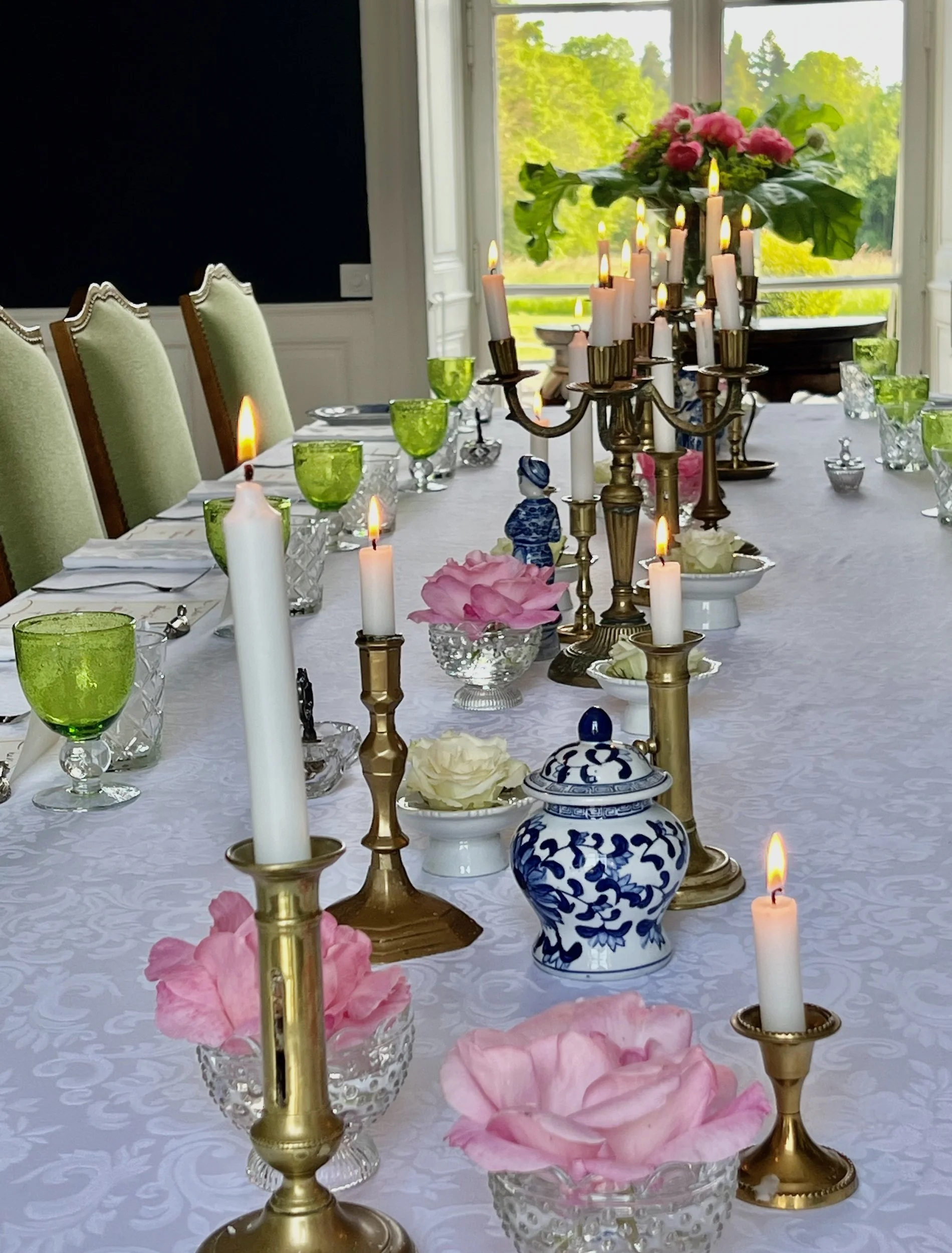 A dining table decorated with candles, pink and white flowers, green and blue glassware, and ornate cutlery, set near a large window with a view of greenery outside.