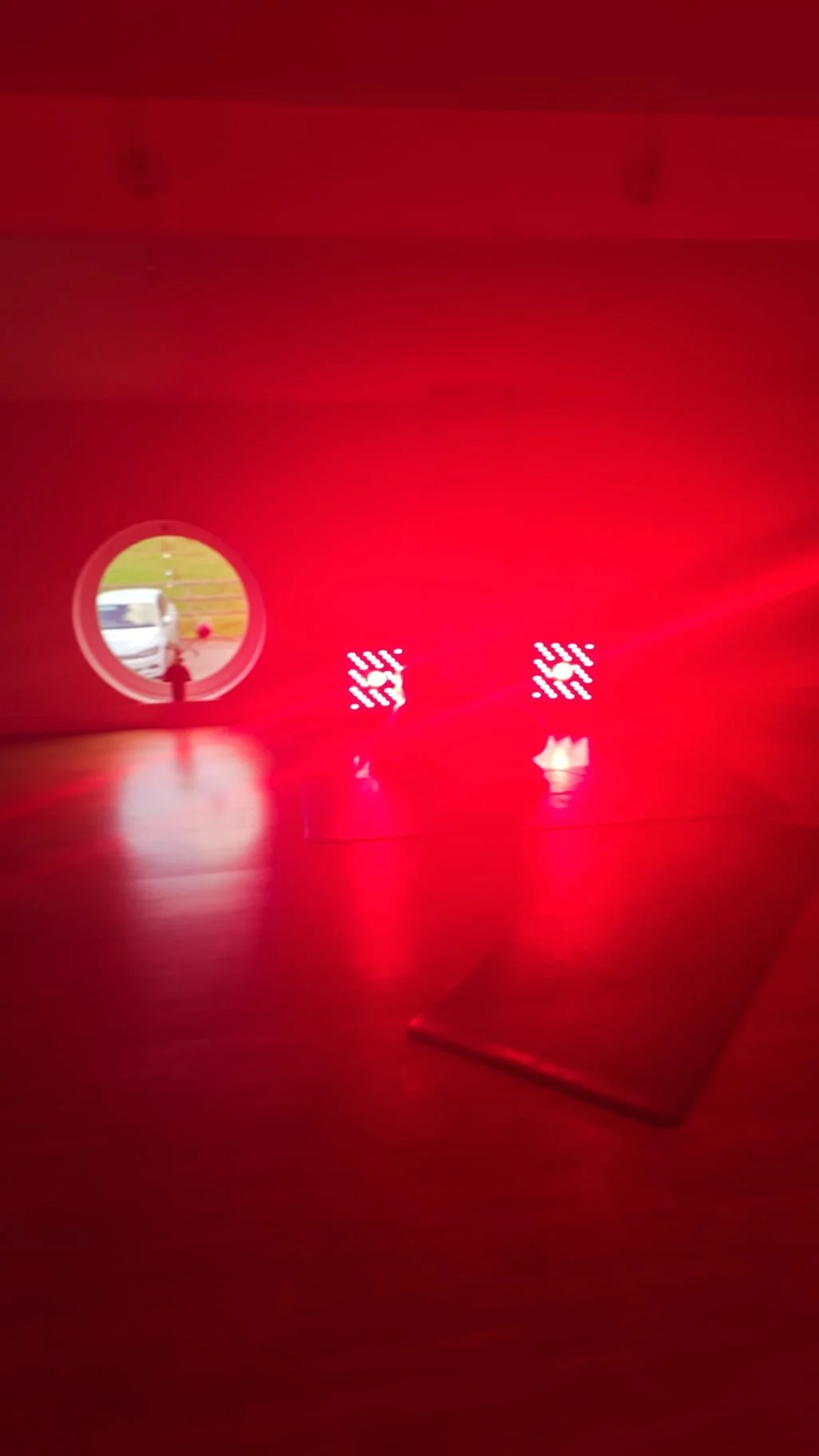 Interior of a room with red lighting, a small circular window showing a person and a vehicle outside, and illuminated decorative panels on the wall.