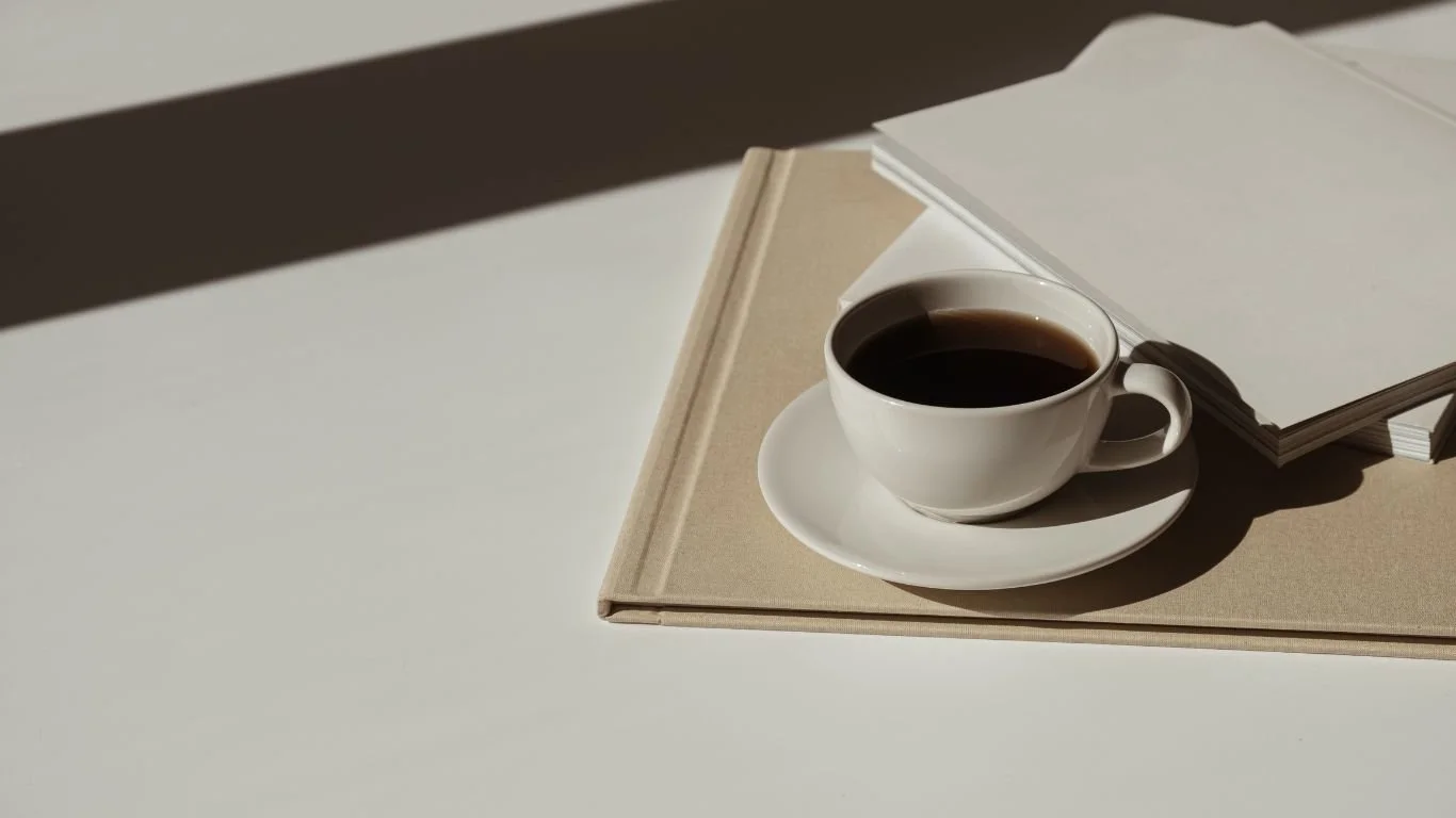 A white coffee cup filled with black coffee on a saucer, placed on top of closed books and notebooks on a white table, casting a shadow in natural light.