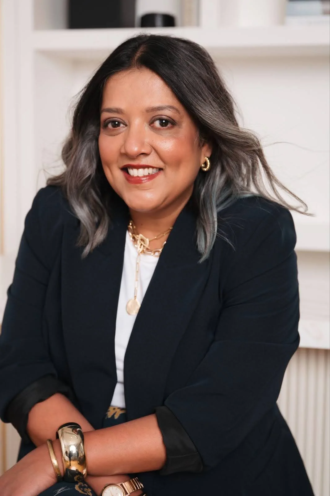 A woman with shoulder-length wavy hair, wearing a black blazer, white top, gold jewelry, and a watch, smiling while sitting in a well-lit room with white walls and shelves.