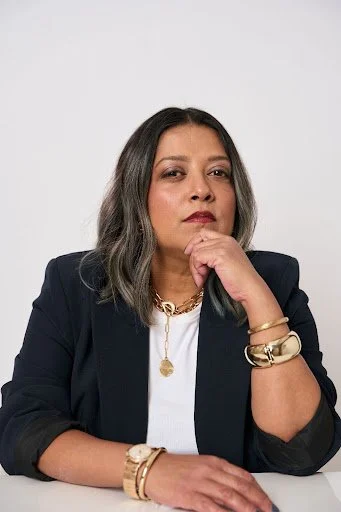 A woman with shoulder-length gray hair, wearing a black blazer over a white top, sitting with her right elbow on a white surface, hand resting near chin, looking confident against a plain white background.