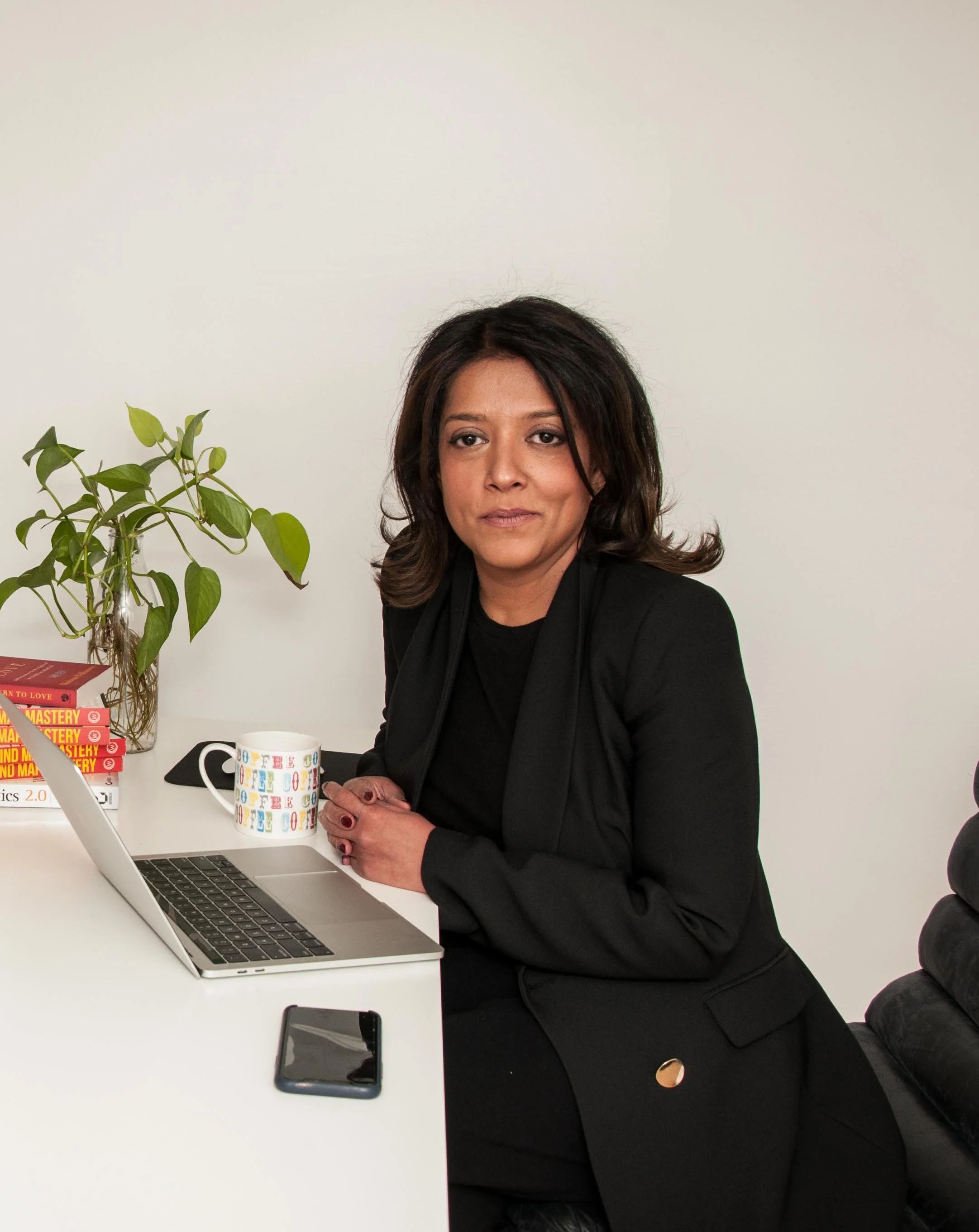 Professional woman sitting at a desk with a laptop, coffee mug, and phone, wearing a black blazer in a minimalist office with a plant and books in the background.