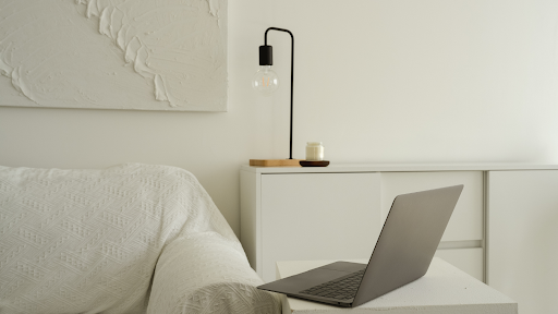A bedroom scene with a white bed, a white dresser, and a wall-mounted lamp, with a laptop on the dresser.