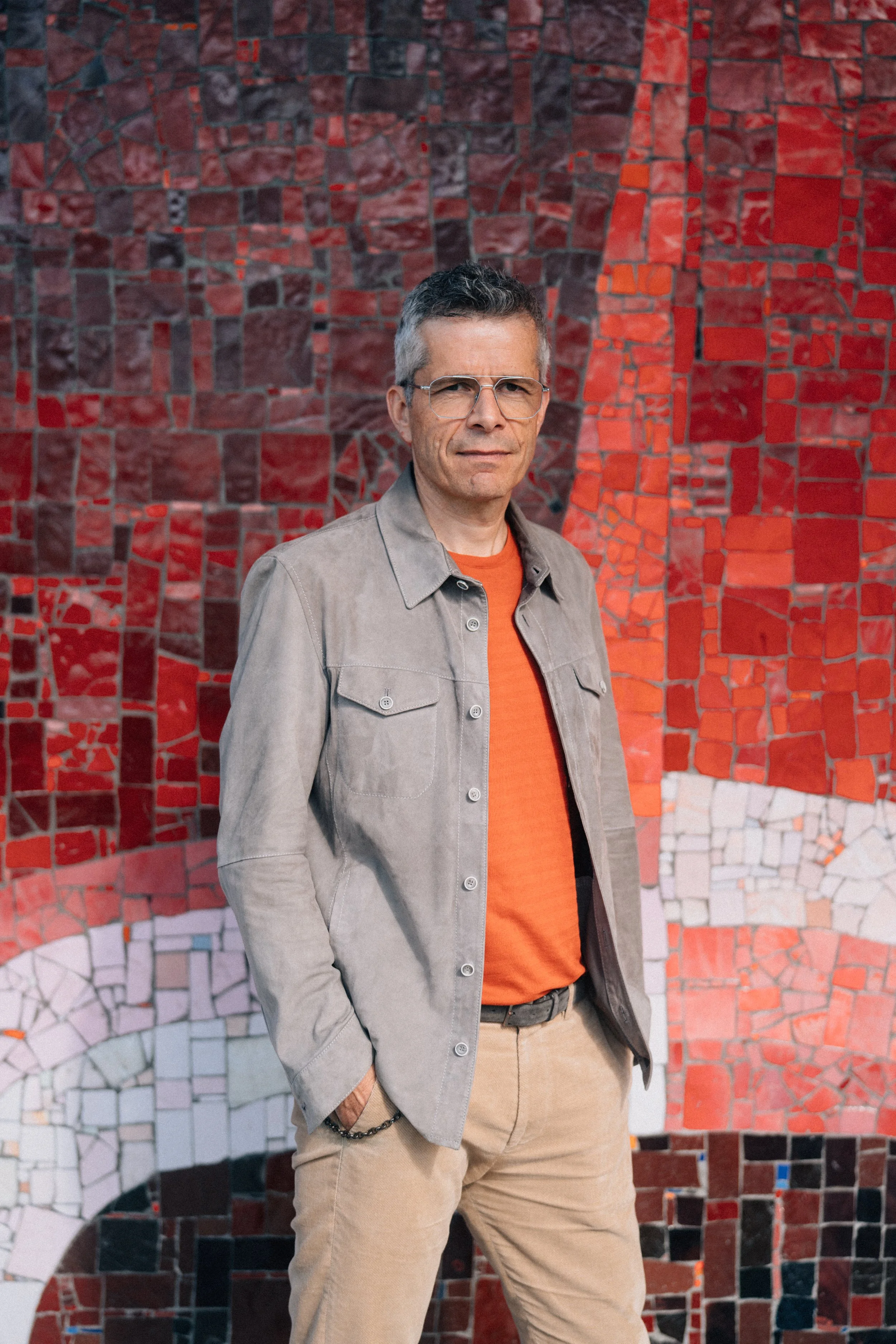 Ein Mann steht vor einer bunten Mosaikwand, trägt eine Brille, ein beige Hemd, ein rotes T-Shirt und beige Hosen.
