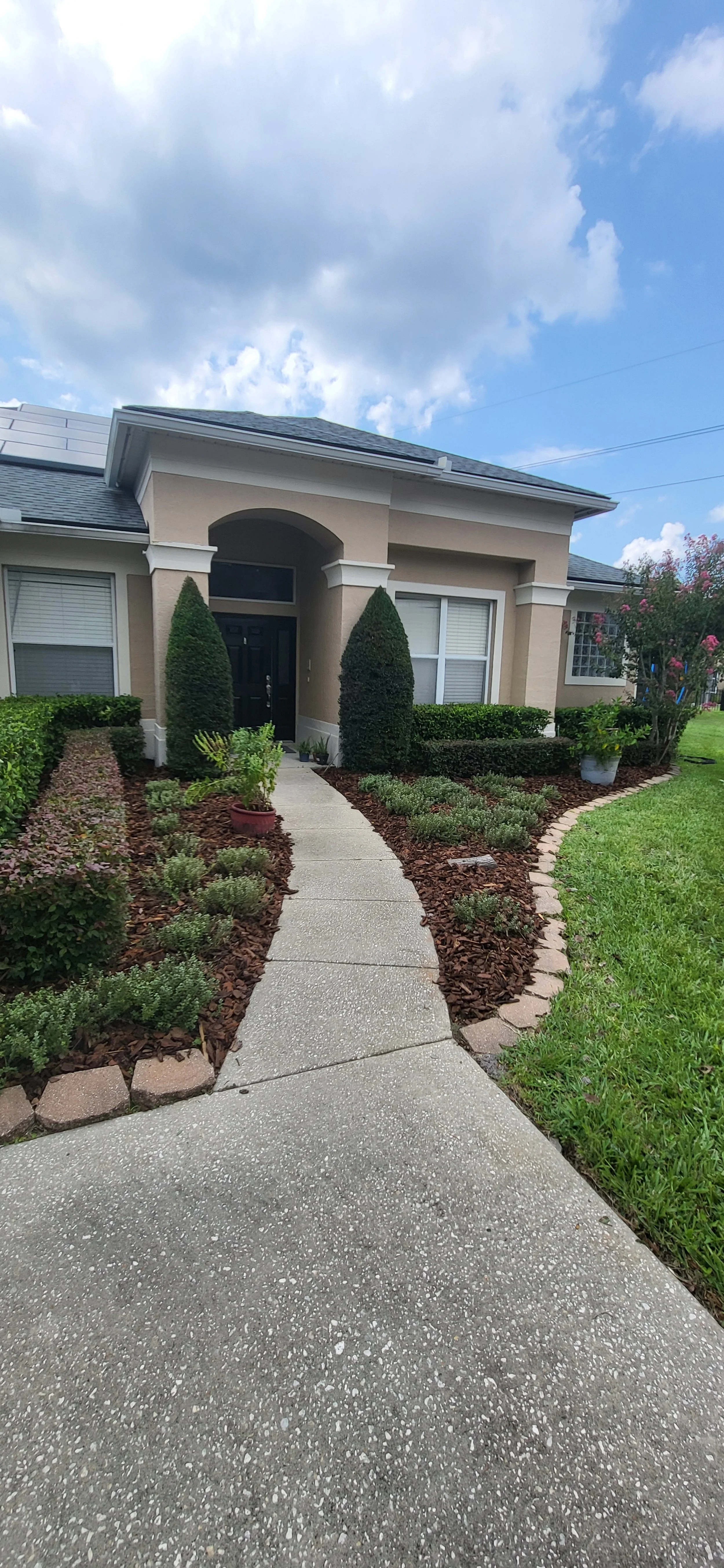 Front view of a house with a curved walkway, manicured bushes, and flowering plants near the entrance, under a partly cloudy sky.