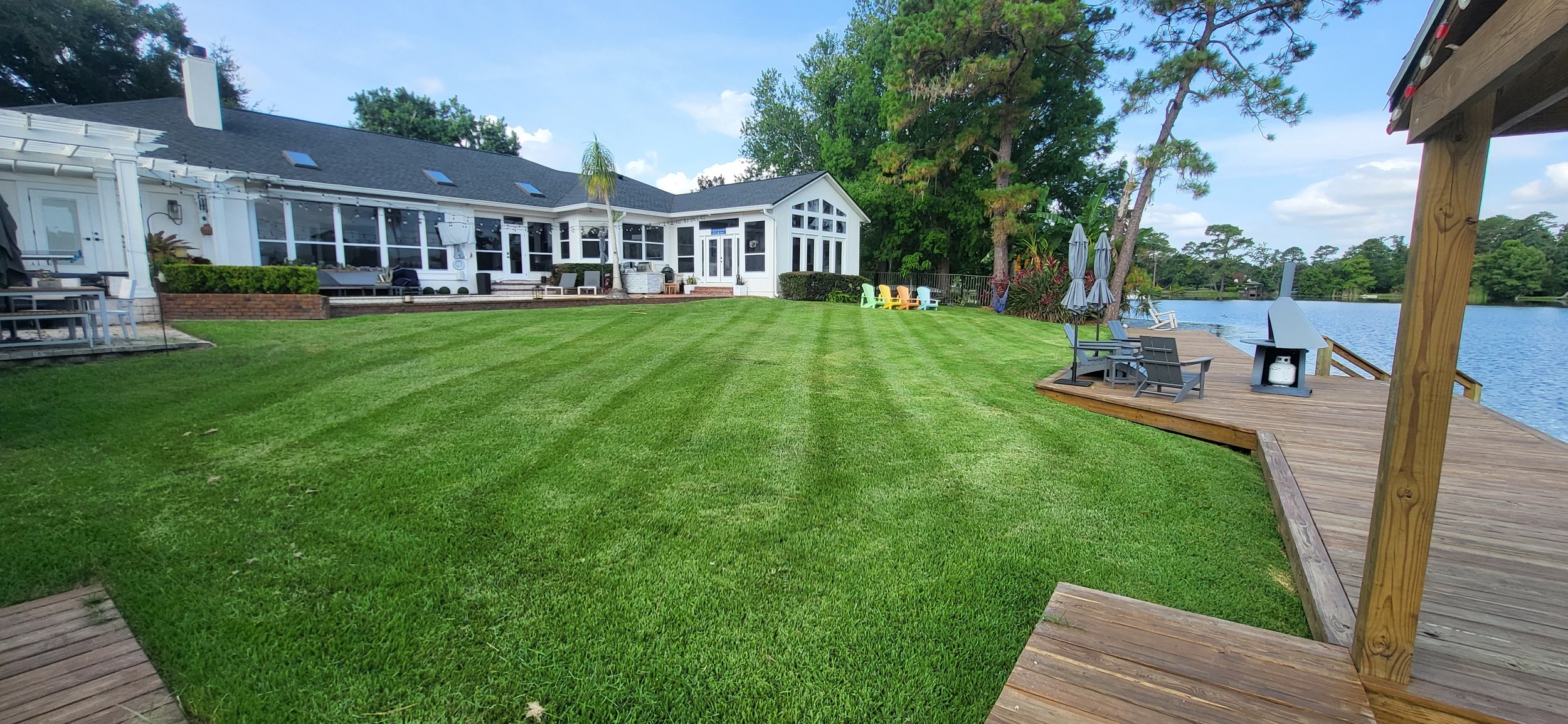 A backyard with a large green lawn, a white house with a glass-enclosed sunroom, a wooden deck with outdoor furniture, and a lake with trees in the background.