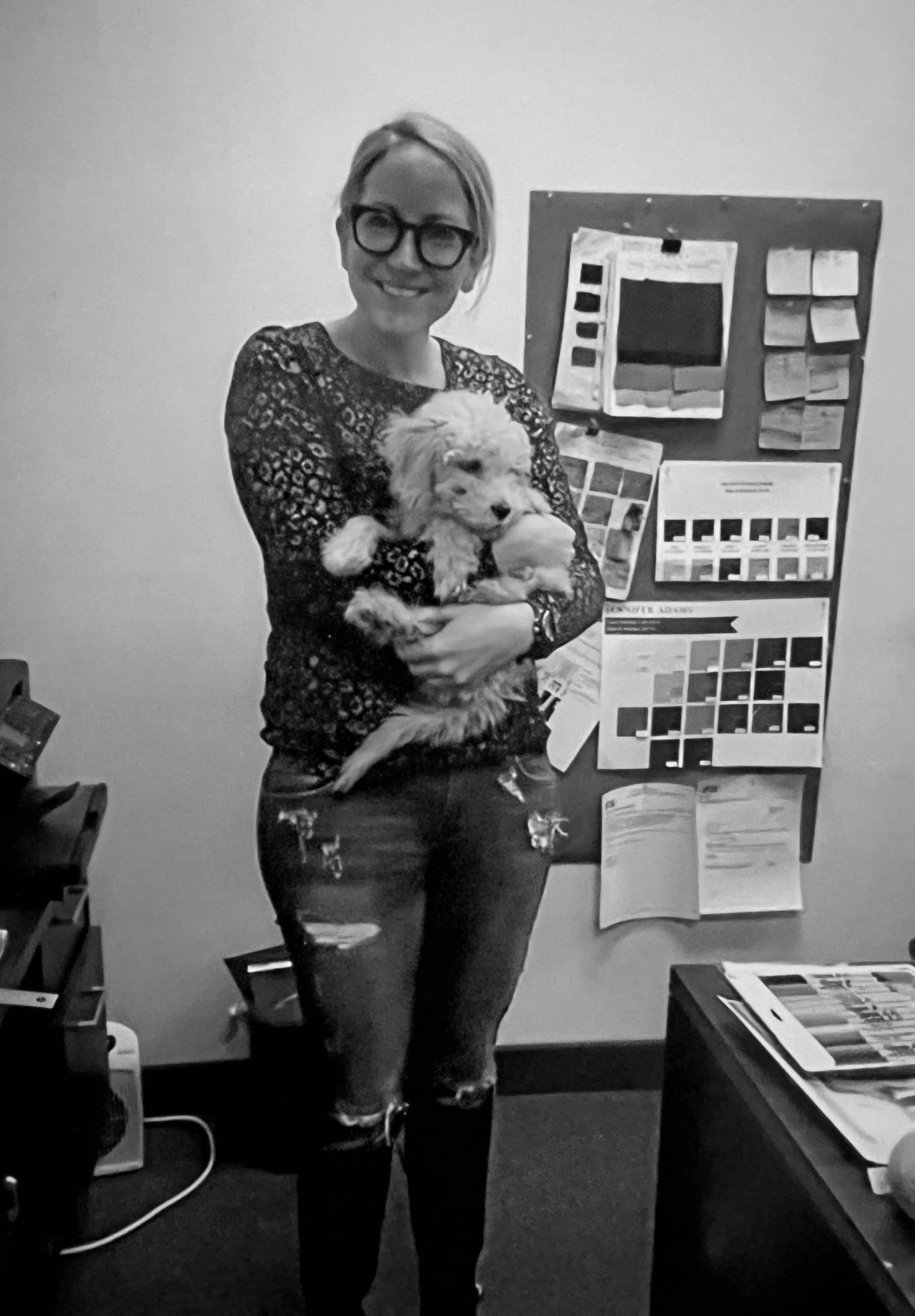 Person holding a small fluffy dog in an office setting with a bulletin board in the background.