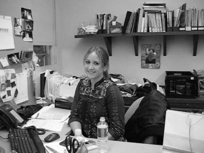 Woman sitting at a desk in an office, surrounded by papers and books, with shelves in the background.