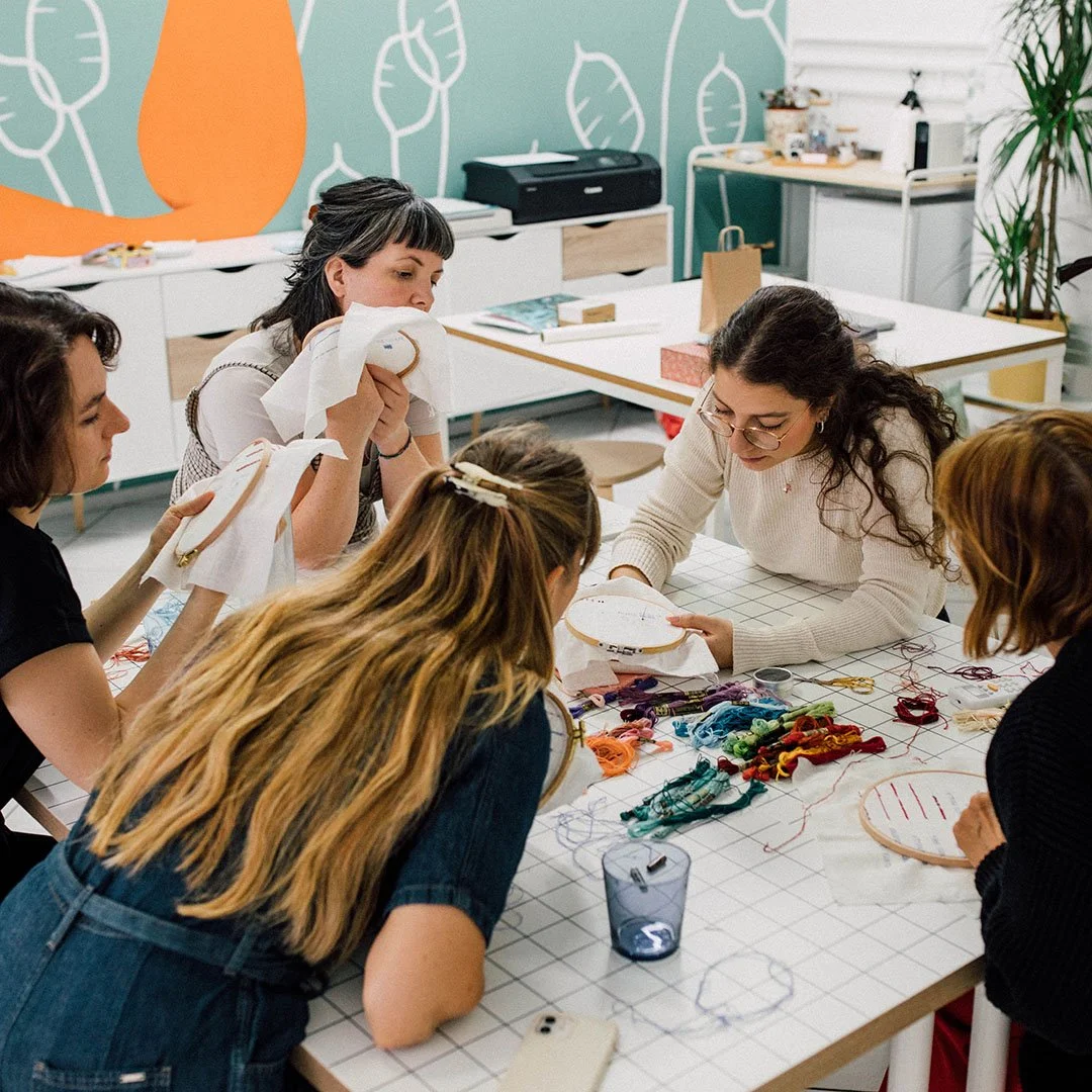 Un gruppo di donne sedute intorno a un tavolo, impegnate in un'attività di ricamo o lavoro a maglia, con fili colorati e telai per ricamo.
