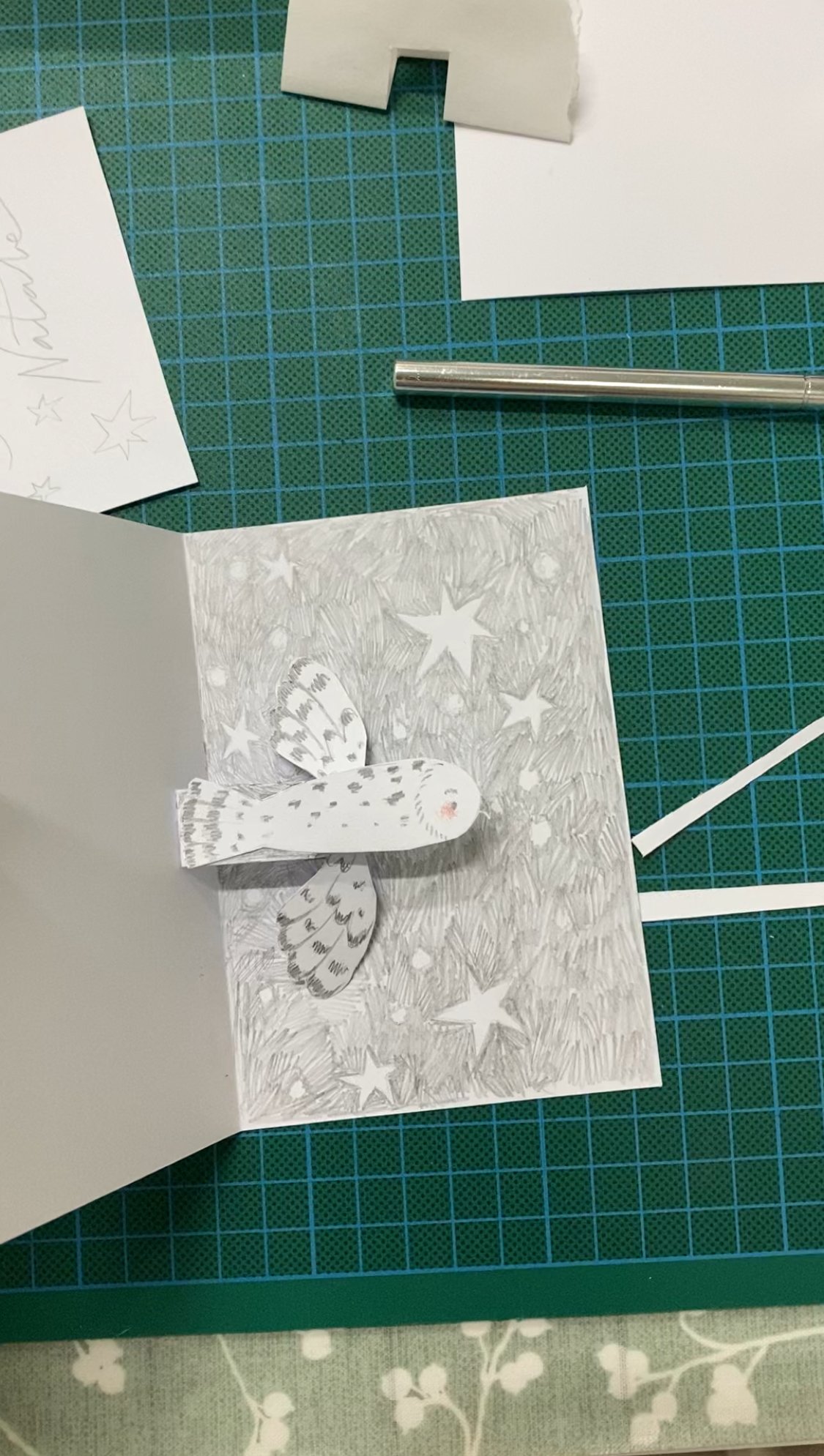 Una cartina di auguri natalizi con una farfalla di carta bianca e dettagli decorativi di stelle e neve, su un tavolo verde con strumenti di carta.
