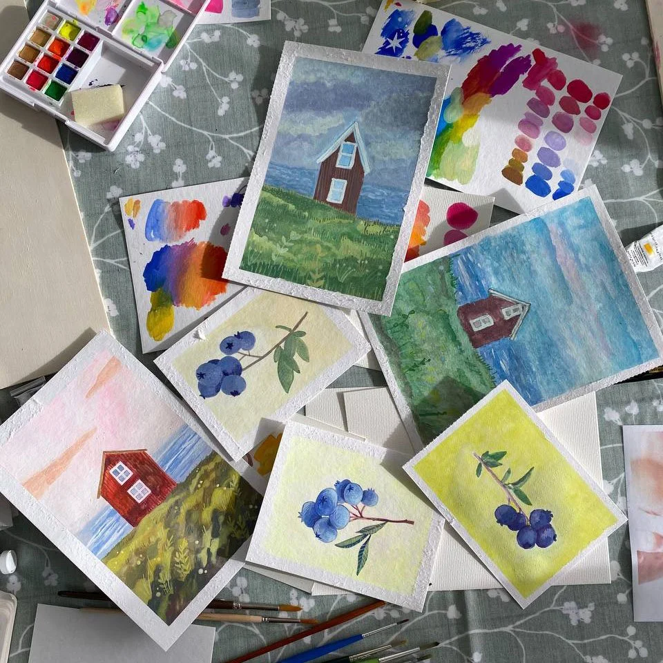Una tavola apparecchiata con vari dipinti ad acquerello di case vicino all'acqua, grappoli di bacche blu e palette di colori.
