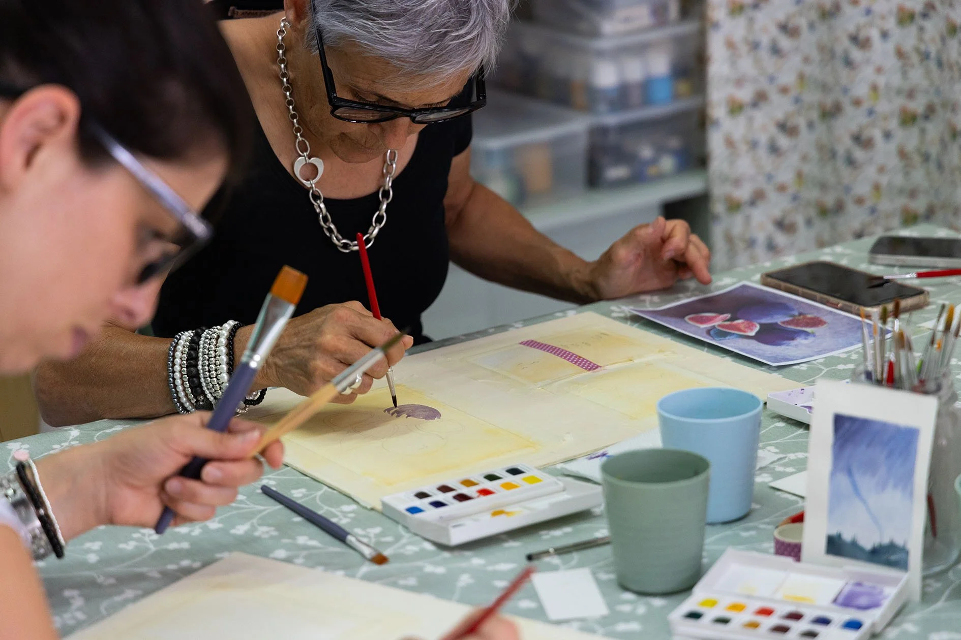 Due donne che dipingono ad acquerello su carta, circondate da colori, pennelli e materiali artistici, in un ambiente creativo.