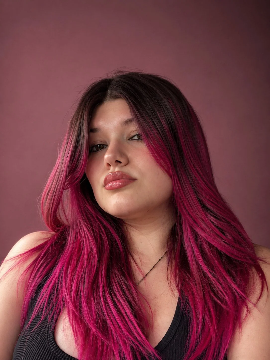 A woman with long, wavy hair that is dark at the roots and bright pink at the ends, looking at the camera with a confident expression, wearing a black top and a delicate necklace, against a muted pink background.