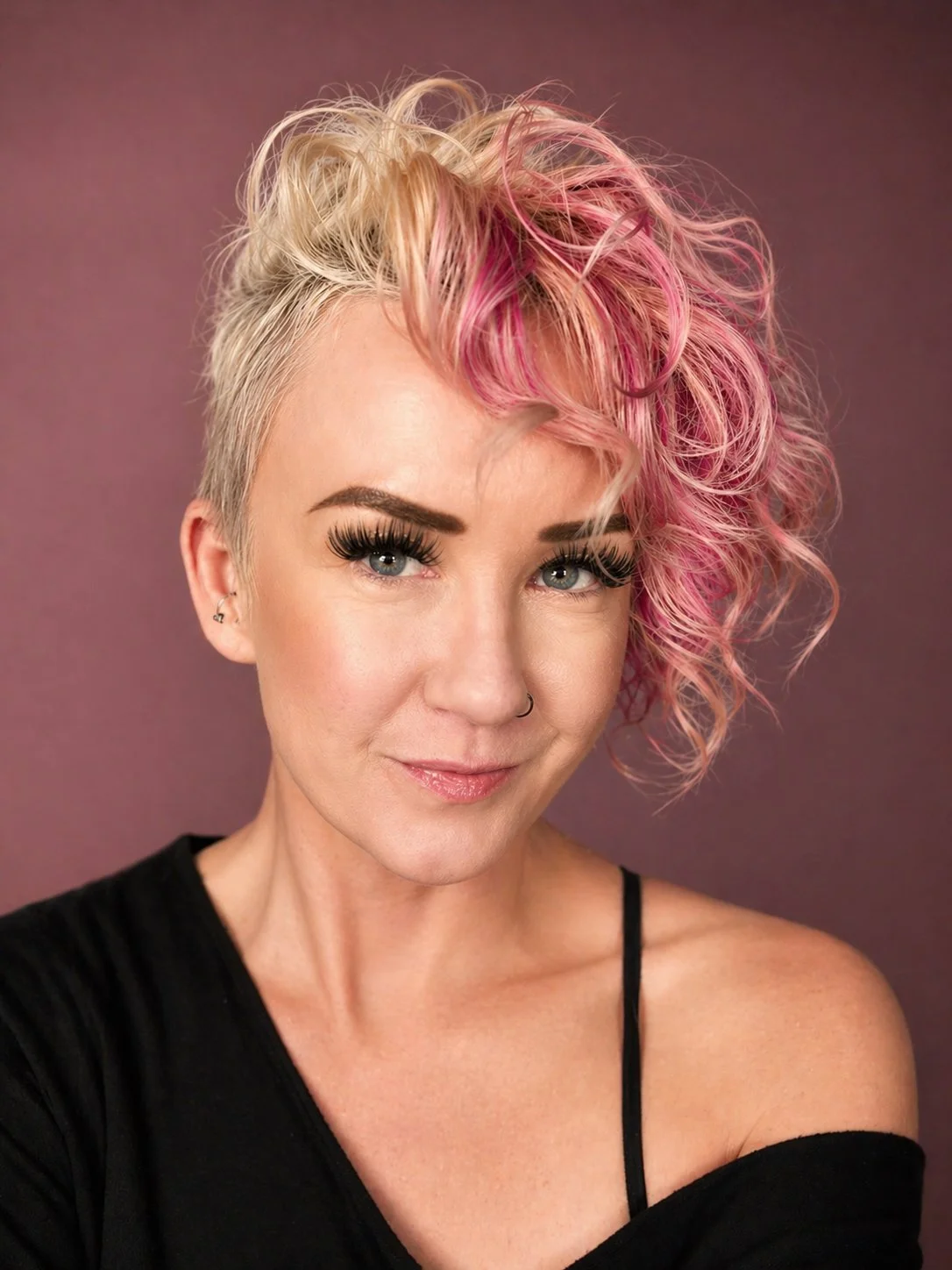 Woman with short blonde and pink curly hair, dark eye makeup, and a nose ring, wearing a black off-shoulder top, against a purple background.